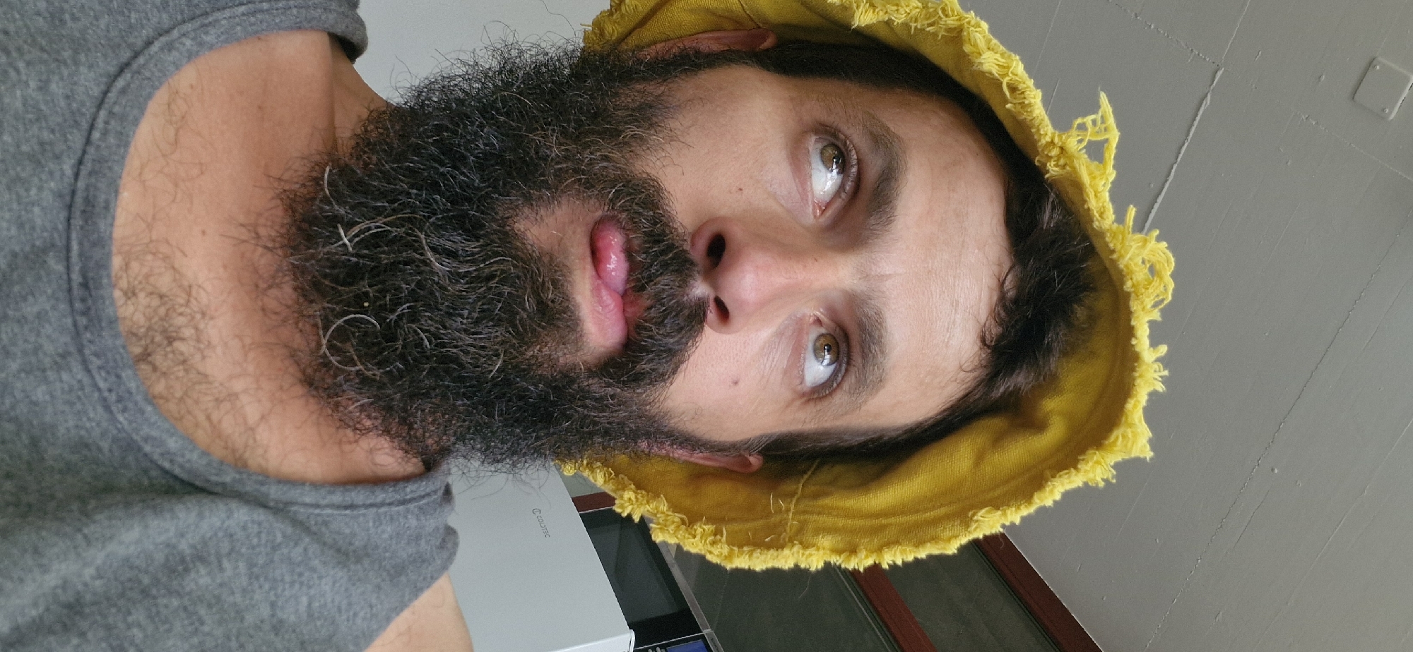 A man with a beard and a yellow hat is sticking his tongue out while looking up and to the side. He is wearing a grey tank top. The man is indoors, with a white ceiling above him. There is a refrigerator behind him, with the brand name "COLDTEC" visible. The image was taken in Uetendorf, Switzerland.