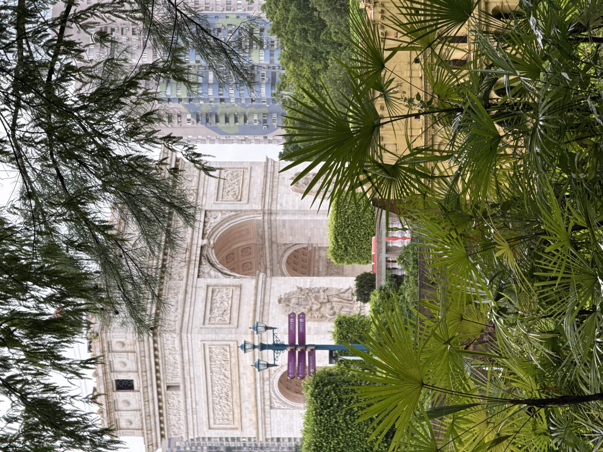 The image depicts a segment of the Window of the World park in Nanshan District, China, showcasing a replica of the Arc de Triomphe. The monument is constructed of white stone and features intricate carvings. In front of the monument, a dark teal lamppost with three lamps stands beside a directional sign with purple plaques. Lush green hedges surround the base of the Arc, and palm trees are prominently featured in the foreground, obscuring parts of the scene. In the background, modern, colorful high-rise buildings rise above the greenery, contrasting with the classical architecture of the monument. The sky is overcast, contributing to a soft, diffused light. There are no people visible in the image.