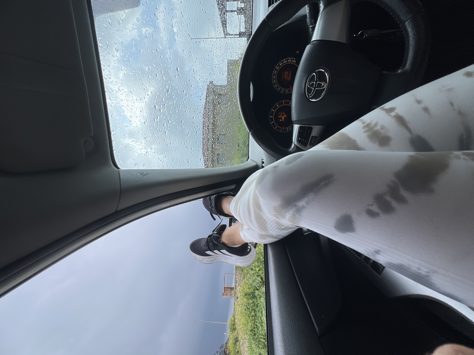 The image depicts an interior view from the driver's seat of a car, presumably stationary. Raindrops are visible on the windshield, obscuring the outdoor view, suggesting inclement weather. The steering wheel and dashboard of a Toyota vehicle are visible. A person's legs, clad in tie-dye leggings, are stretched out across the dashboard with their feet resting against the edge of the open driver's side window. The person is wearing black and white Adidas sneakers. Outside the open window, there is a glimpse of greenery, and in the distance, a small, rudimentary structure is visible. Based on the presence of the structure the scene appears to be located in the city of Prijevor, Montenegro.