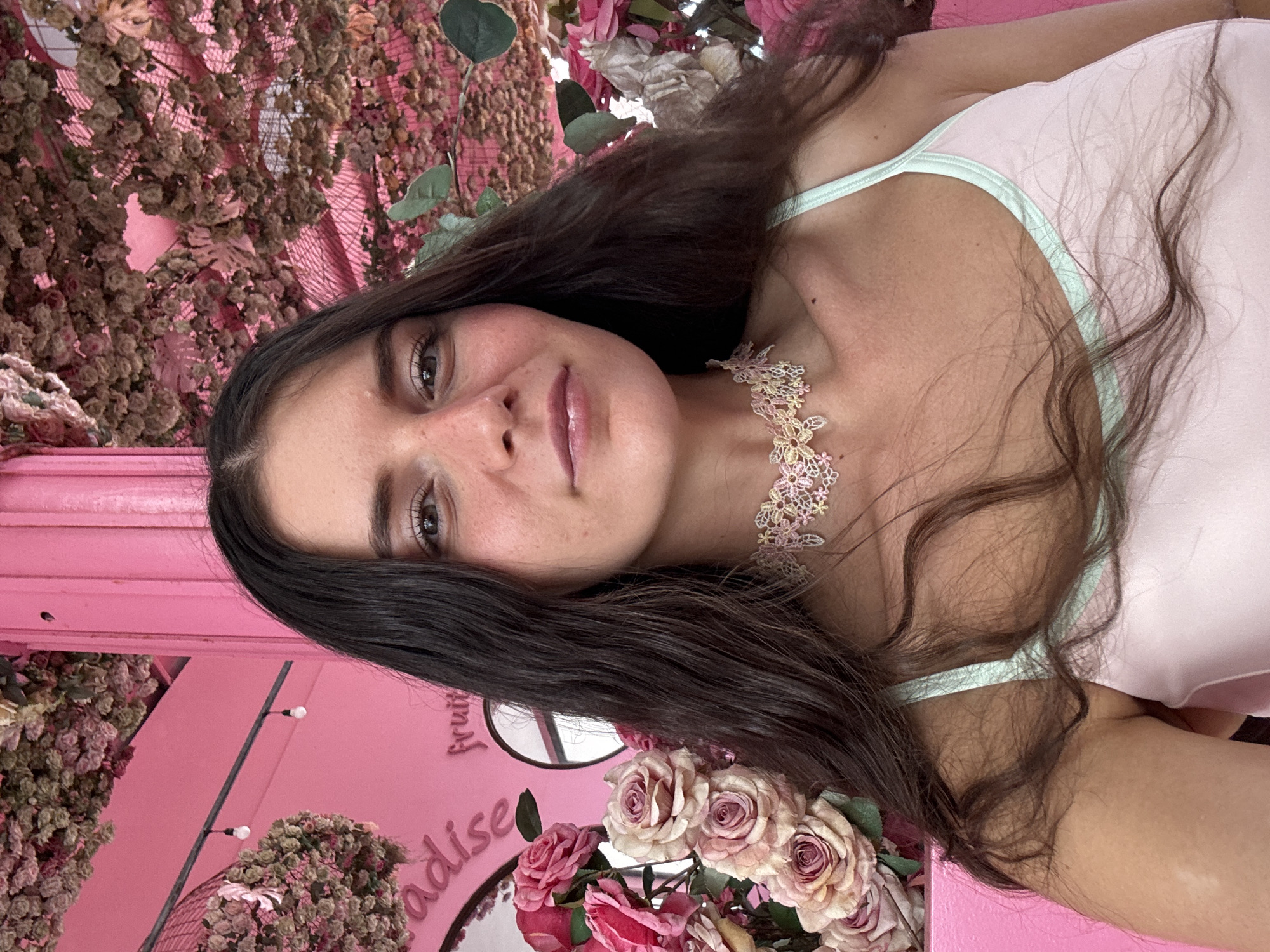 A young woman with long, dark hair and freckles on her face is captured in a selfie. She is wearing a light pink camisole with a mint green trim and a delicate floral choker necklace. The background is vibrantly decorated with pink floral arrangements hanging from the ceiling and on the walls, creating a whimsical atmosphere. A pink column is visible to the left. The scene suggests a playful and aesthetically pleasing environment, possibly a cafe or a themed establishment. The location is identified as Tien Thanh Ward, Vietnam, though no specific landmarks within the ward are visible.