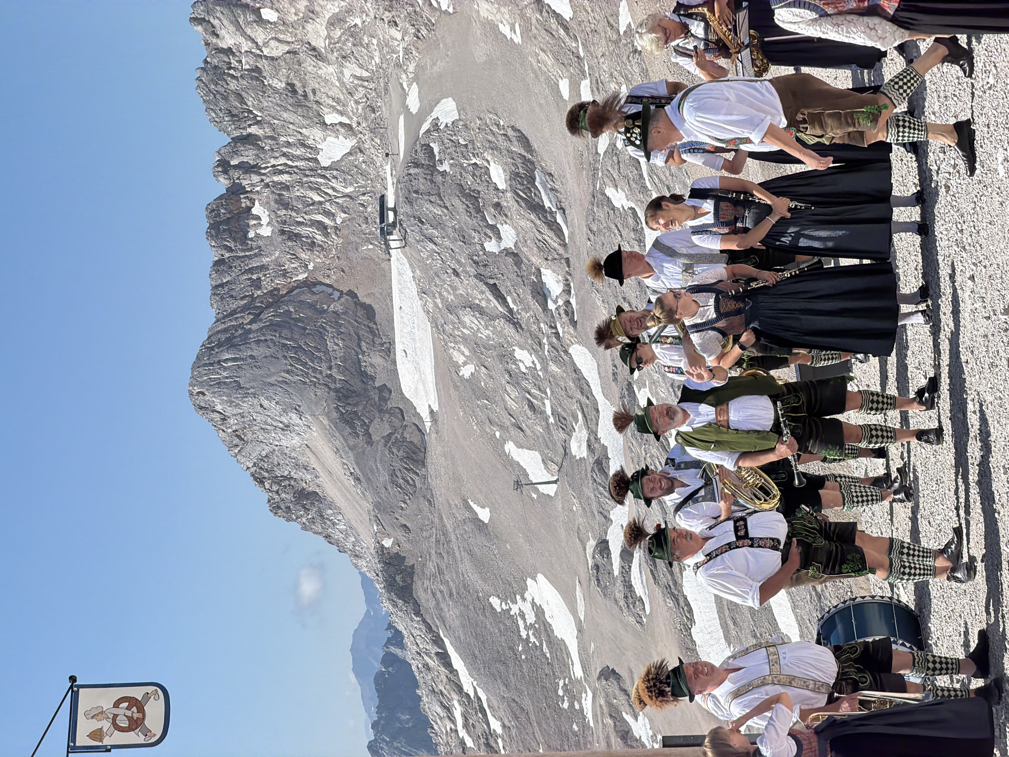 A group of people in traditional Bavarian attire, including Lederhosen and Dirndls, are gathered outdoors on a rocky, mountainous terrain. Several individuals are holding musical instruments such as tubas, clarinets, and a saxophone, suggesting they are a band or ensemble. The background features a large, imposing mountain range with patches of snow, under a clear blue sky. A ski lift structure is visible on the mountain slope. To the left, a pennant with an illustration of a person holding a pretzel hangs from a pole. The people are standing and interacting, with some looking at each other and others looking towards the camera or off into the distance. The overall scene depicts a cultural event or performance in a high-altitude, mountainous setting.