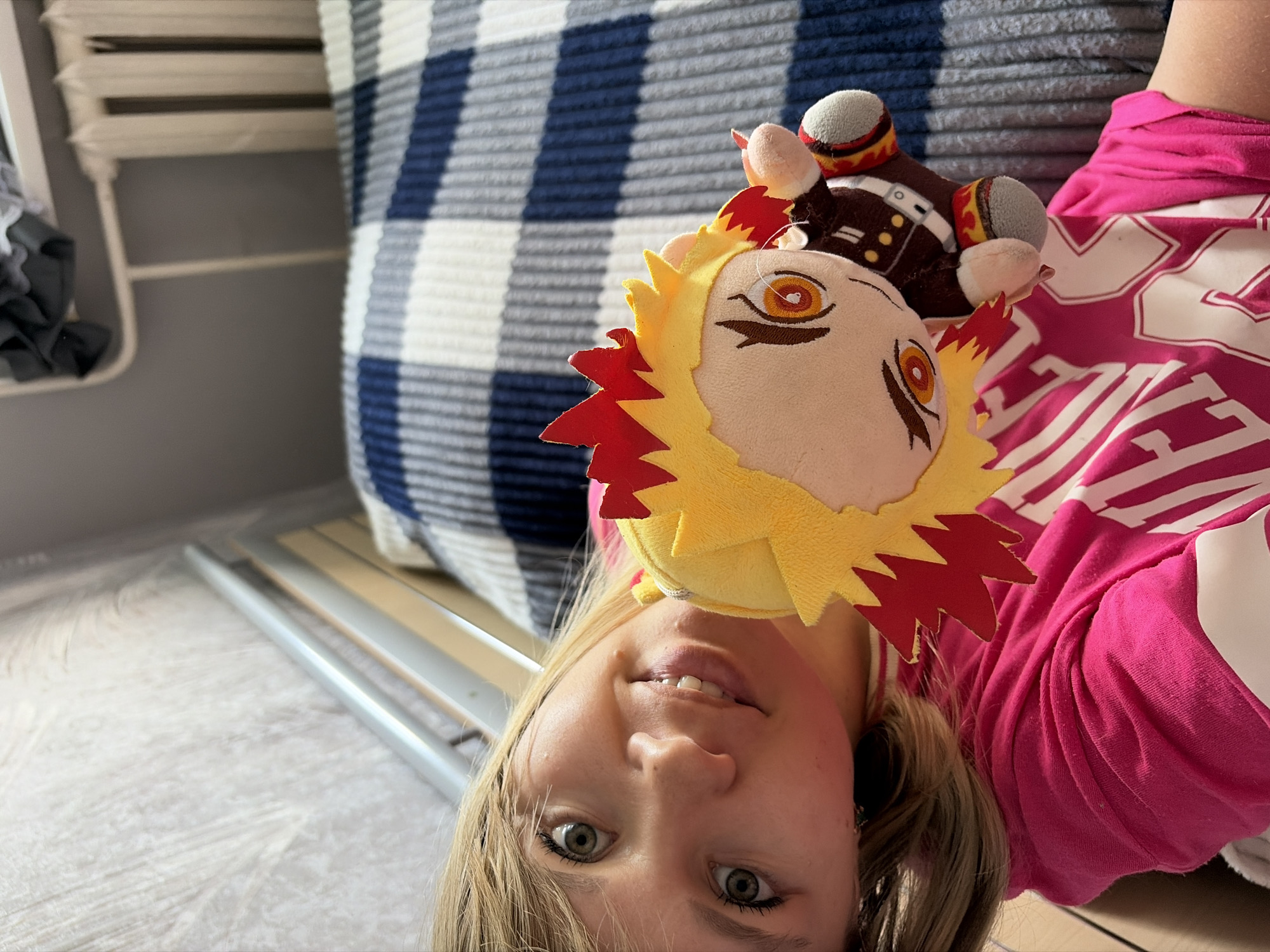 A young woman is holding a Rengoku Kyoujurou plush doll. She is wearing a pink shirt with white stripes. The background includes a blue and white striped pillow. The location is in a room with a white metal frame visible on the floor. The city is Chaikovsky, Russia.