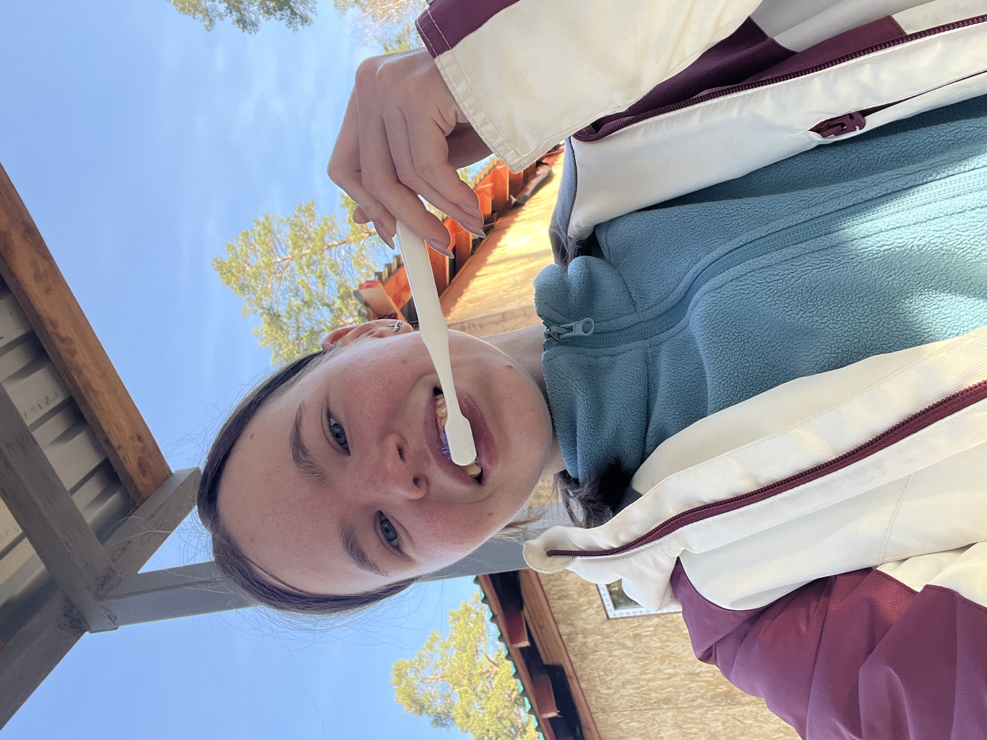 A young woman is holding a toothbrush in her mouth. She is wearing a blue fleece jacket under a white and burgundy windbreaker. The background shows trees and a partially visible wooden building with a dark red roof. A portion of a metal structure is visible on the left. Based on the location provided, this scene is likely to be located in Lesnoye, Russia.