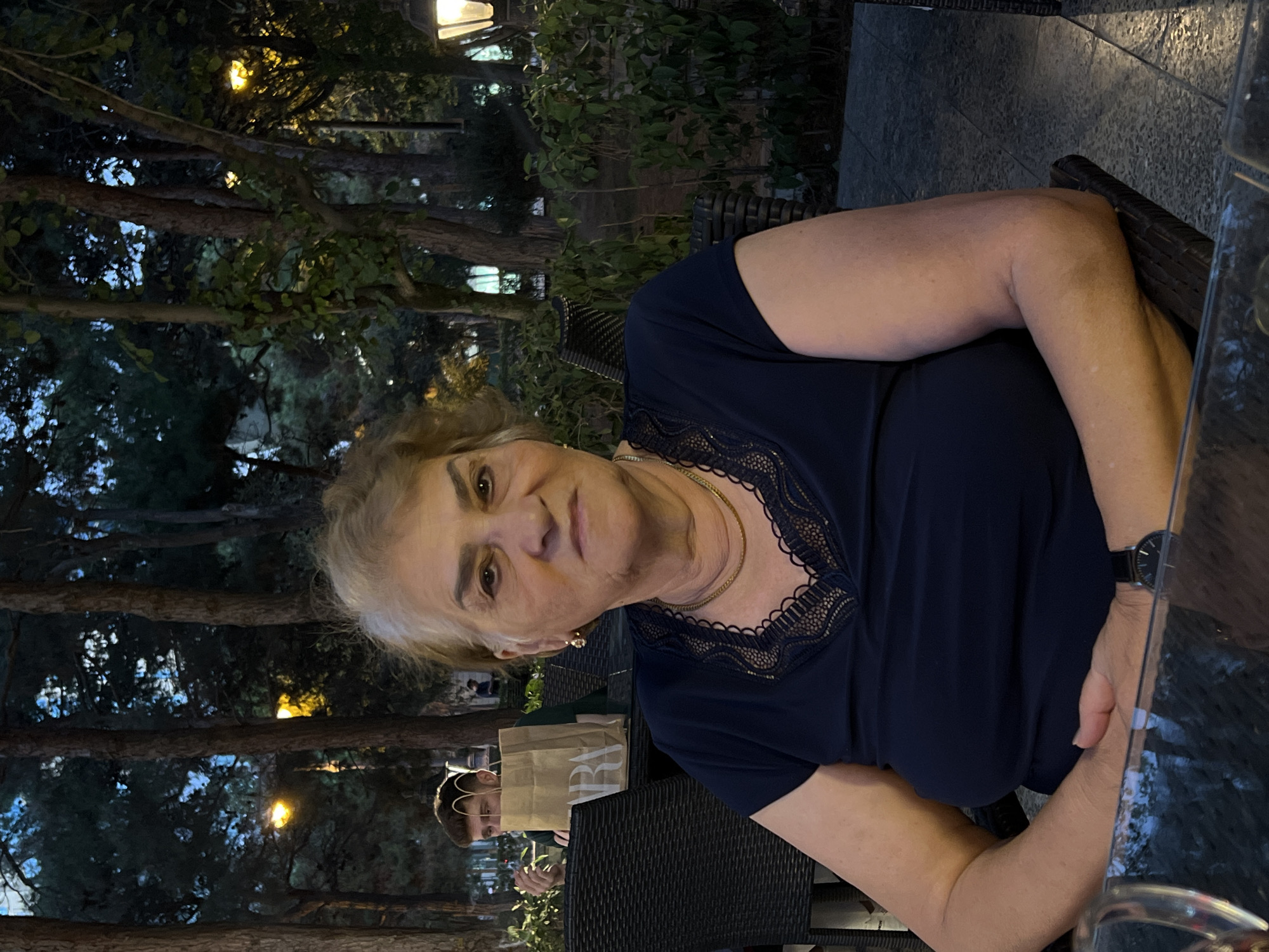 The image shows an elderly woman seated at a table outdoors in what appears to be a park or restaurant setting. She is looking directly at the camera with a neutral expression. She is wearing a dark blue top with lace detailing around the neckline and a gold necklace. A watch is visible on her left wrist. Behind her, a young man is visible in the background, partially obscured by a paper shopping bag. He is looking away from the camera and appears to be holding something to his mouth. The environment is characterized by trees, foliage, and outdoor lighting, suggesting an evening or dusk setting. The presence of tables and chairs indicates a social or dining area. The location is identified as Sabail Raion, Azerbaijan.