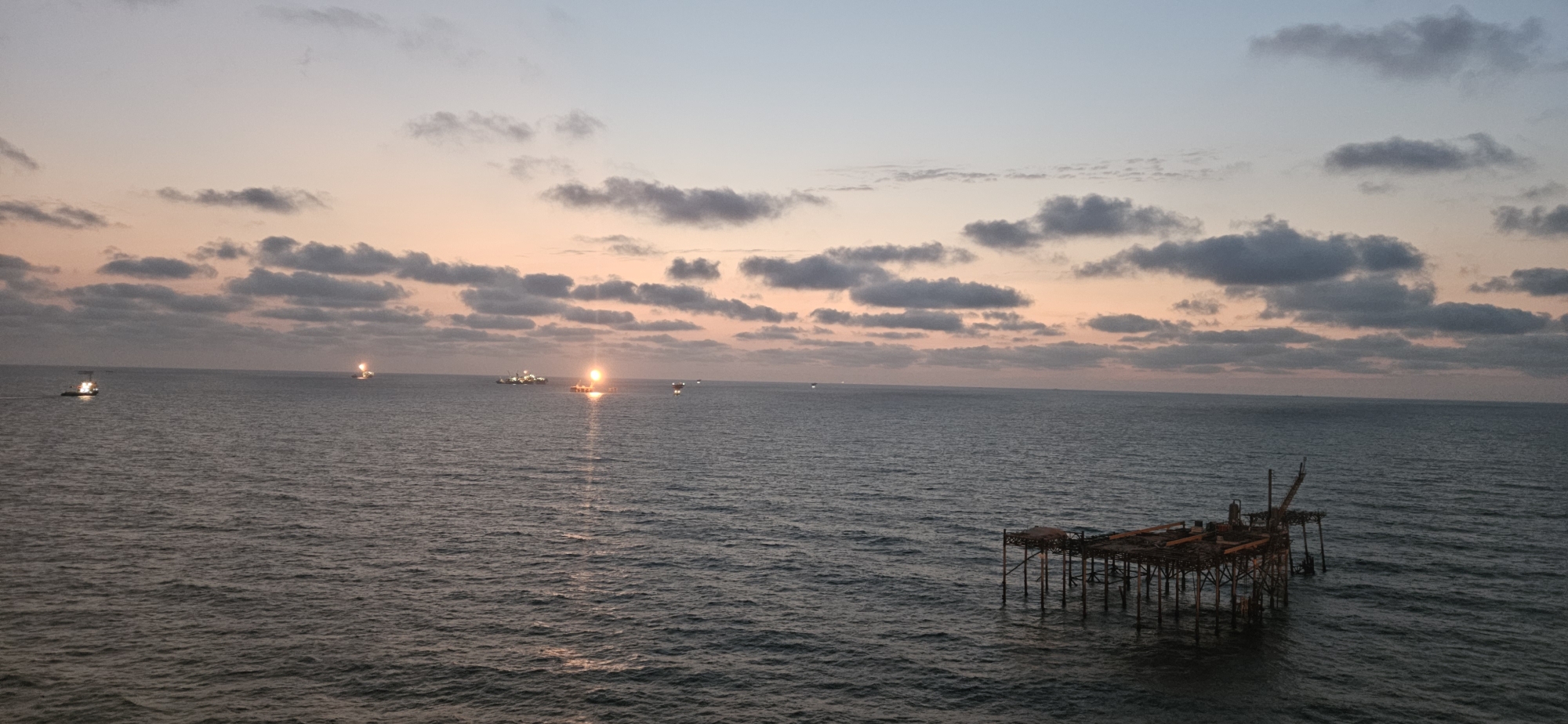 The image depicts a panoramic view of the ocean at dusk. The sky is painted with hues of pink, orange, and pale blue, with scattered dark clouds. Several lights are visible across the water, indicating vessels or structures. In the foreground, to the right, is a partially constructed or abandoned wooden pier extending into the sea. The water is calm with small waves. The overall scene suggests an offshore industrial or marine environment.
