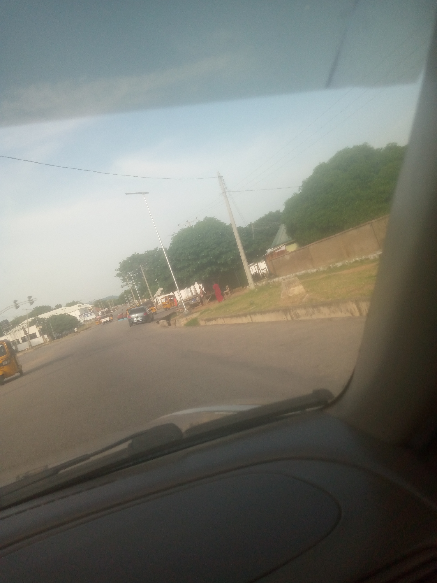 The image depicts a street scene in Jalingo, Nigeria, viewed from inside a vehicle. The road is paved and appears to be a main thoroughfare. Several vehicles are visible, including a yellow tricycle (likely a 'Keke Napep'), cars, and a larger truck. The time of day seems to be late afternoon or early evening, with soft, diffused light and a hazy sky, possibly due to dust or cloud cover.
On the sides of the road, there are buildings, some with white facades, and a long, beige-colored wall. Lush green trees and vegetation are abundant, suggesting a tropical or sub-tropical climate. A prominent street light stands on a tall pole. The presence of traffic lights at an intersection further indicates this is an urban area with traffic management. The overall atmosphere appears calm, with no obvious signs of distress or urgency. The image captures everyday life in Jalingo, with people and vehicles going about their business.
