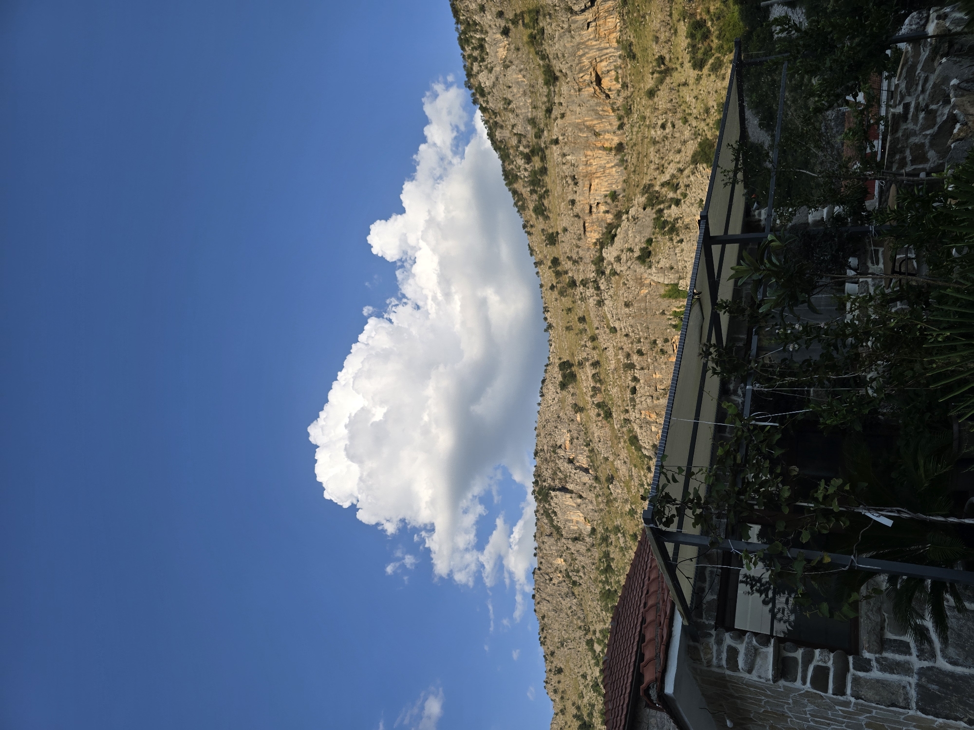 In Podgorica, Montenegro, a clear blue sky with a prominent, fluffy white cloud is visible above a rugged, sun-drenched mountain. The mountain slopes are covered with sparse vegetation and rocky outcrops. In the foreground, a stone house with a red-tiled roof is partially visible, featuring a covered patio area with plants. The scene captures a serene outdoor environment.