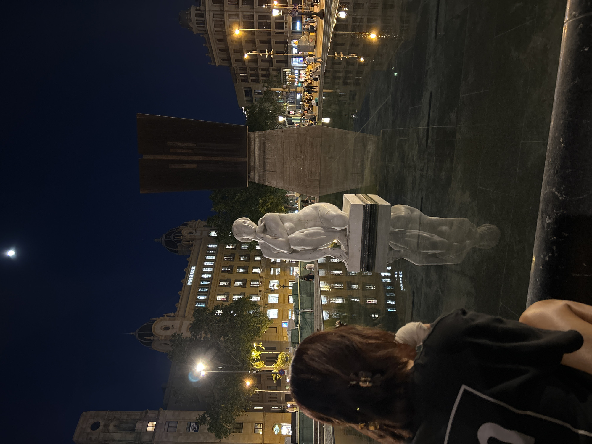 The image depicts a night scene in Barcelona, Spain, likely in Plaça Catalunya. In the foreground, a person is sitting, with their back partially visible, facing a fountain or reflective surface. The fountain reflects a statue of a person in a reclining pose. Behind the statue and fountain, there are buildings, one of which has the word "PRIMARK" illuminated. The sky is dark, with a crescent moon visible. The area is lit by streetlights, and there are people walking in the background, reflected in the water. A modern art sculpture stands near the statue.