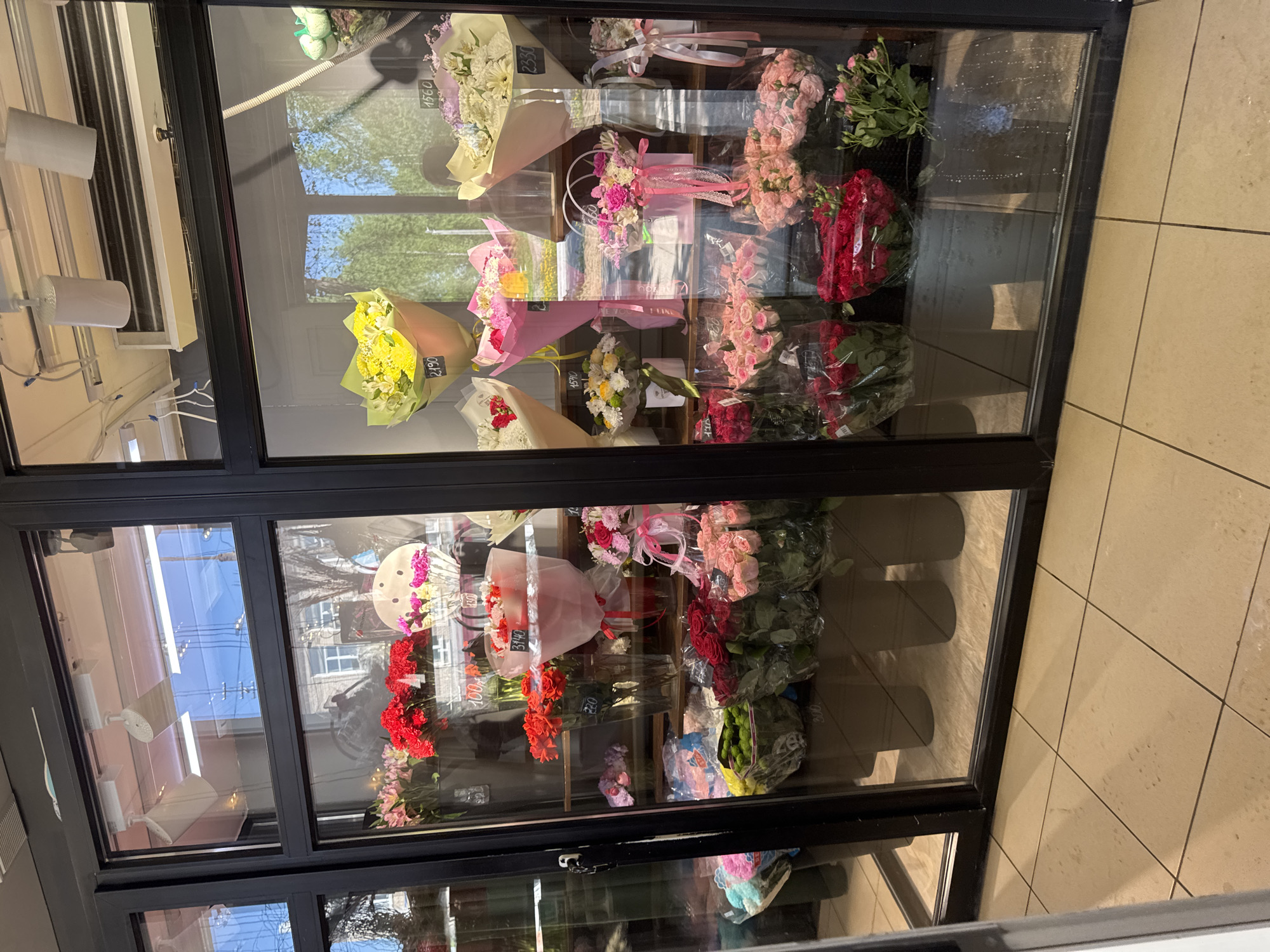 A photograph captures the inside of a floral display case, likely located in the city of Chaikovsky, Russia. The case features multiple bouquets of flowers in various colors and arrangements, including roses, chrysanthemums, and other species. Some bouquets are wrapped in paper or cellophane, and price tags are visible on some. The display is behind glass panels with black frames. Reflections are visible on the glass, showing the street outside. The floor tiles in front of the display are beige.