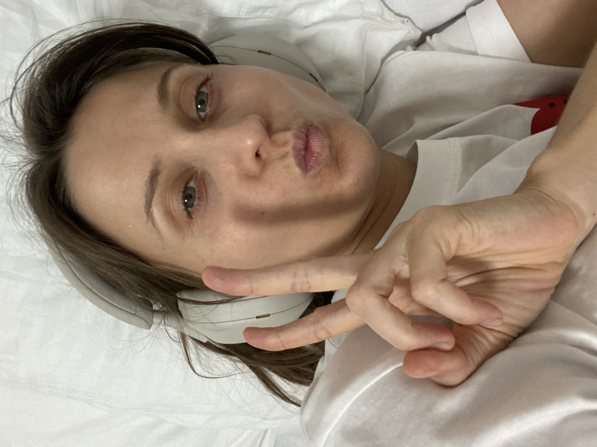 A young woman is captured in a close-up, high-angle selfie, lying on white bedding indoors. She has dark brown hair and light-colored eyes, looking directly at the camera with a playful expression. Her lips are pouted in a "duck face," and her right hand is raised to form a peace sign. She is wearing white over-ear headphones and a white t-shirt, which reveals a hint of a red design or pattern peeking from beneath its folds. Her left hand is also visible, resting on the crumpled white bedding. The setting appears to be a bed in an indoor environment, creating a casual and relaxed atmosphere. This scene is located in Budva, Montenegro. No visible text is clearly discernible.
