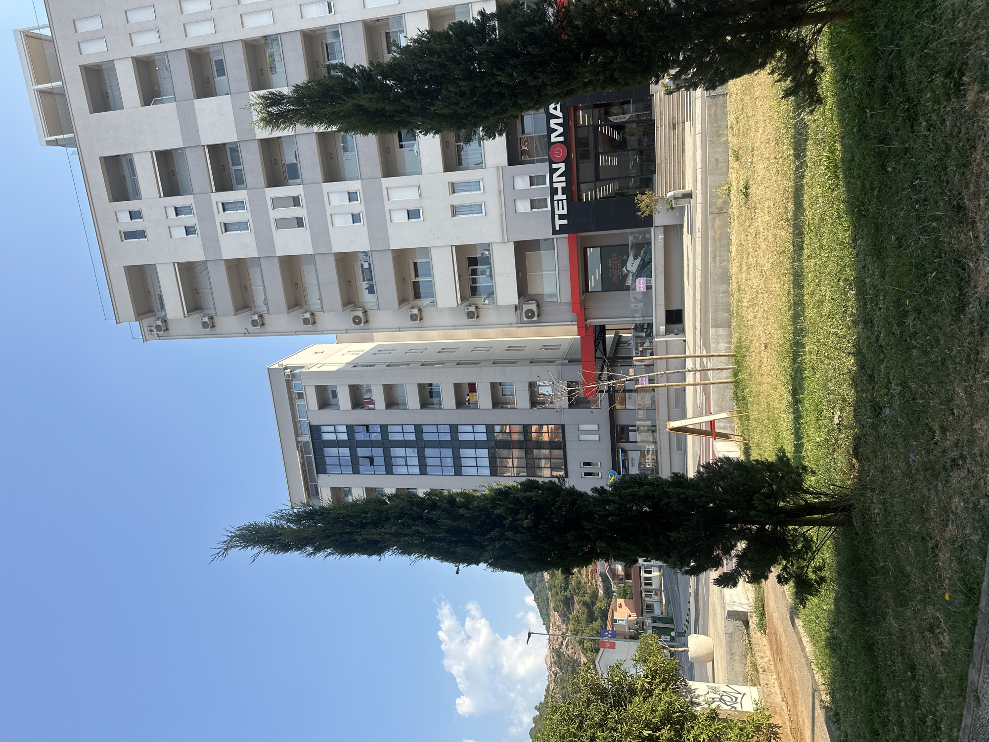 In Budva, Montenegro, on a bright, clear day, two modern apartment buildings stand tall. The building on the right is taller and wider, featuring a grey facade with balconies and numerous windows. The building on the left is narrower and slightly shorter, also with a grey facade and a section of dark-tinted windows. In the foreground, two tall, slender evergreen trees are positioned in a grassy area, with a paved walkway and a street visible beyond. A store with a large sign that reads "TEHNOMAX" is situated at the base of the right building, its entrance accessible by a set of stairs. Further back, more buildings, a street with a few cars, and a hillside covered in vegetation and rocks are visible. The sky is a clear, vibrant blue with a few scattered white clouds.