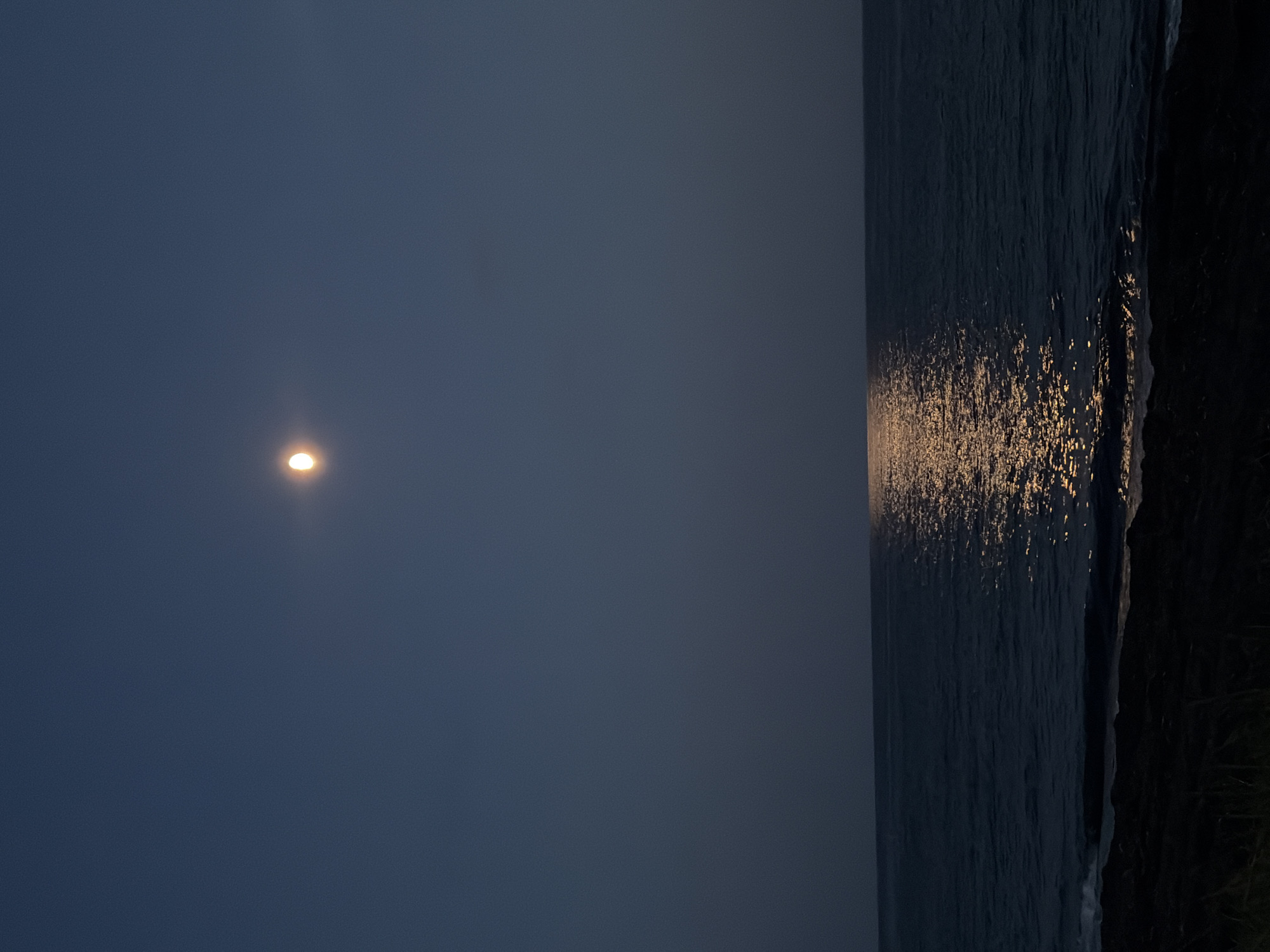 The image depicts a dark night scene at a body of water, likely a sea or large lake, with a prominent full moon illuminating the sky. The moon is positioned in the upper center of the frame, casting a bright reflection on the water's surface that stretches towards the viewer. The water itself appears calm with gentle waves lapping at a dark, rocky shore in the foreground. The sky is a deep, inky blue, transitioning to black towards the edges. There is no visible evidence of human activity, structures, or any specific landmarks that would indicate the location within the city of Ozerskoe, Russia. The overall atmosphere is serene and tranquil, dominated by the natural elements of the moonlit night.