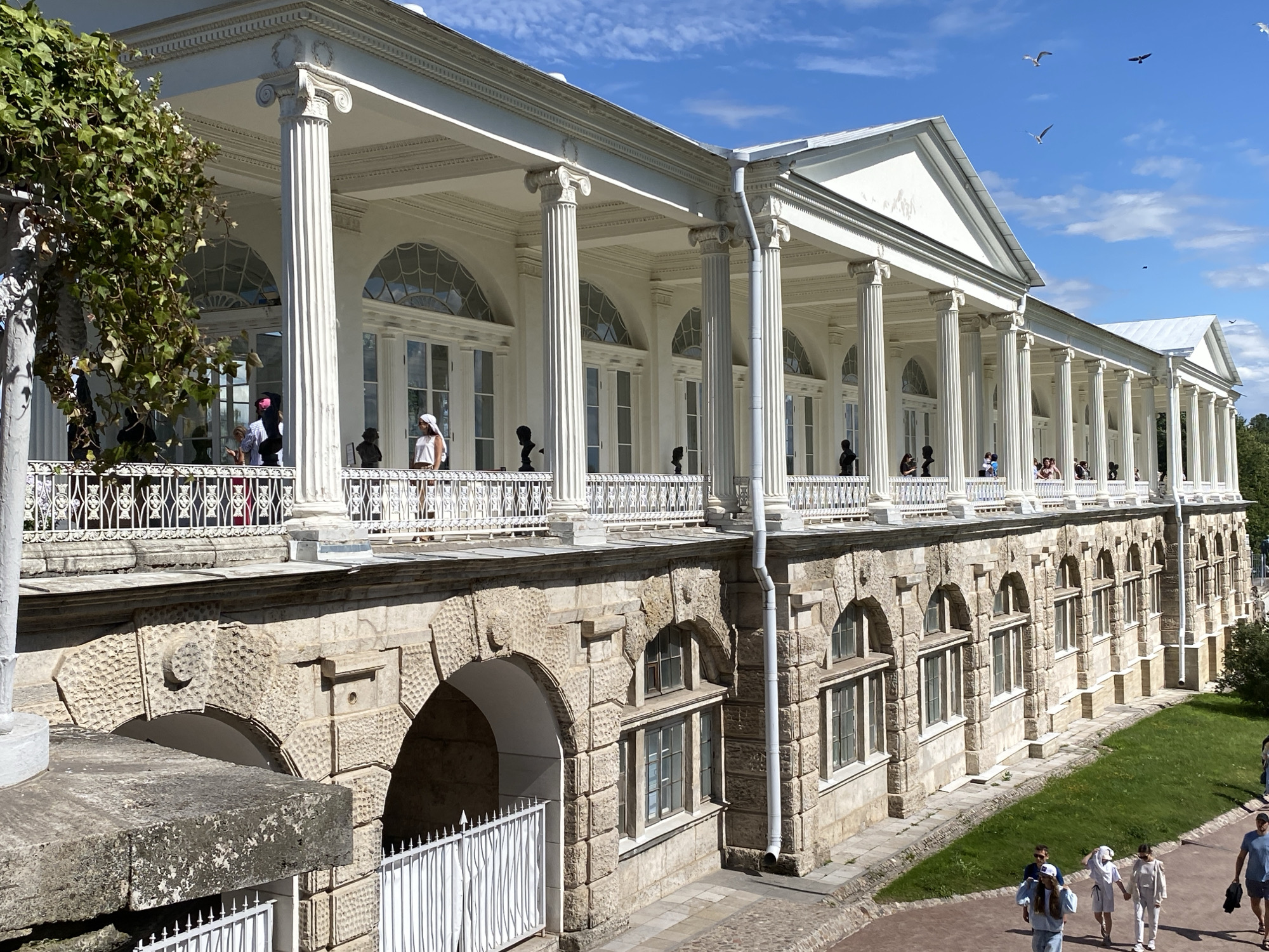 The image depicts the Catherine Palace, also known as the "Agate Rooms," in Pushkin, Russia, on a sunny day. The palace features a long, white colonnade with Ionic columns and a white balustrade. In front of the colonnade, there are several people, some standing and observing, and others walking. The lower level of the palace is constructed of rough-hewn stone with arched openings and windows. A white metal fence is visible in front of some of the arched openings on the lower level. Beyond the palace, a green lawn slopes away, and trees are visible in the distance. The sky is blue with scattered white clouds, and several birds are flying overhead. The overall scene conveys a sense of architectural grandeur and a pleasant outdoor atmosphere.