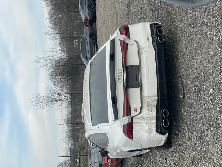 A white Audi S7 Sportback is observed from the rear, parked on a coarse gravel lot in Voynegovtsi, Bulgaria. The vehicle is dirty, with pronounced vertical dirt streaks on the rear bumper and trunk. The right rear taillight assembly is missing its outer lens, exposing internal components, and white tape is affixed to the remaining light structure, the upper right rear window edge, and the adjacent roofline. A dent is visible on the white rear bumper below the right-center parking sensor. The left rear taillight is intact. The vehicle features quad exhaust tips and a black license plate frame devoid of a license plate. To the immediate left, a portion of a red vehicle is visible. In the background, two dark BMW sedans are parked on a similar gravel surface. A green metal fence, topped with coiled razor wire, separates the vehicle area from a dense line of bare deciduous trees and bushes. Faint industrial structures are discernible beyond the vegetation under an overcast sky. No individuals or active interactions are present in the scene.