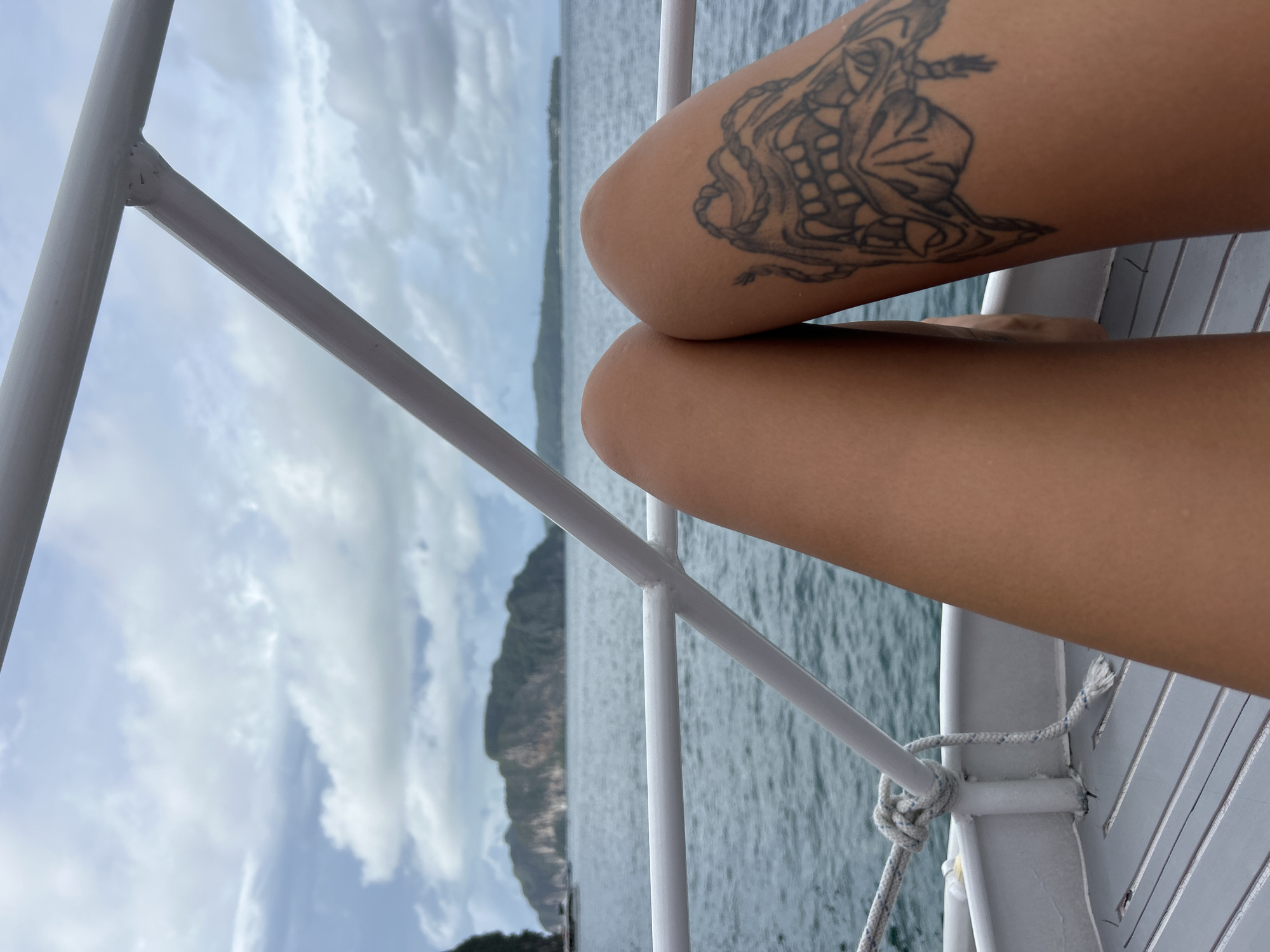 A person's legs are visible in the foreground of the image, with a tattoo on the right thigh. The person appears to be sitting on a boat deck, with the railing of the boat in the middle ground. In the background, there is the ocean and a large island. The sky is cloudy. The scene is likely in Ao Nang, Thailand, given the presence of the distinctive limestone karst islands common to the region, which are a major tourist attraction.