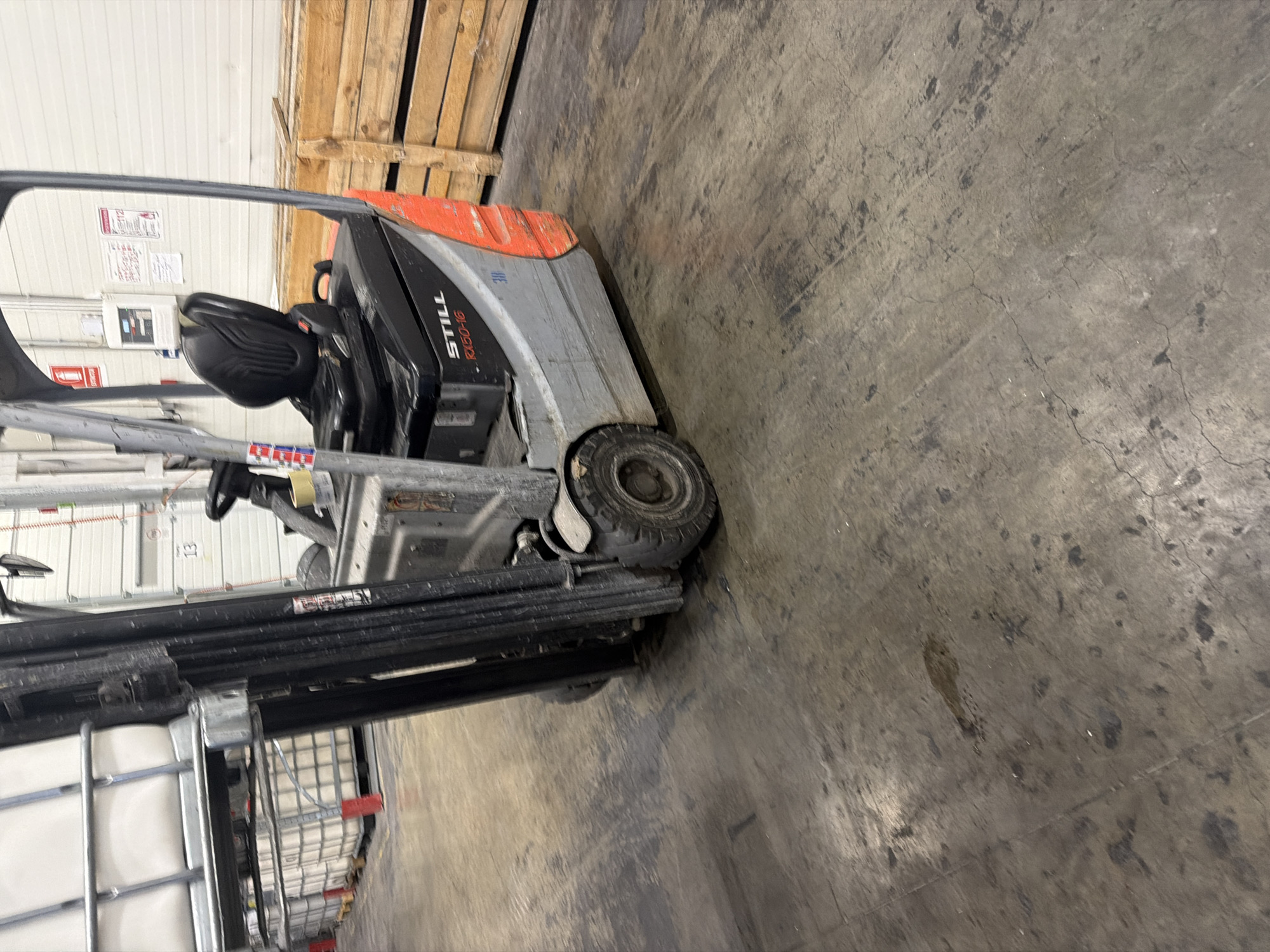 An indoor warehouse scene shows a forklift positioned on a concrete floor. The forklift is primarily gray and orange, with a black seat and steering wheel. The brand "STILL RX50-16" is visible on the side of the forklift. Behind the forklift, stacks of wooden crates are present, and in the background, white garage doors are visible. Further in the distance, numerous large white containers are stacked. The concrete floor exhibits signs of wear, including stains and cracks. The overall environment suggests an operational warehouse or storage facility. The image was captured in Strășeni, Moldova.