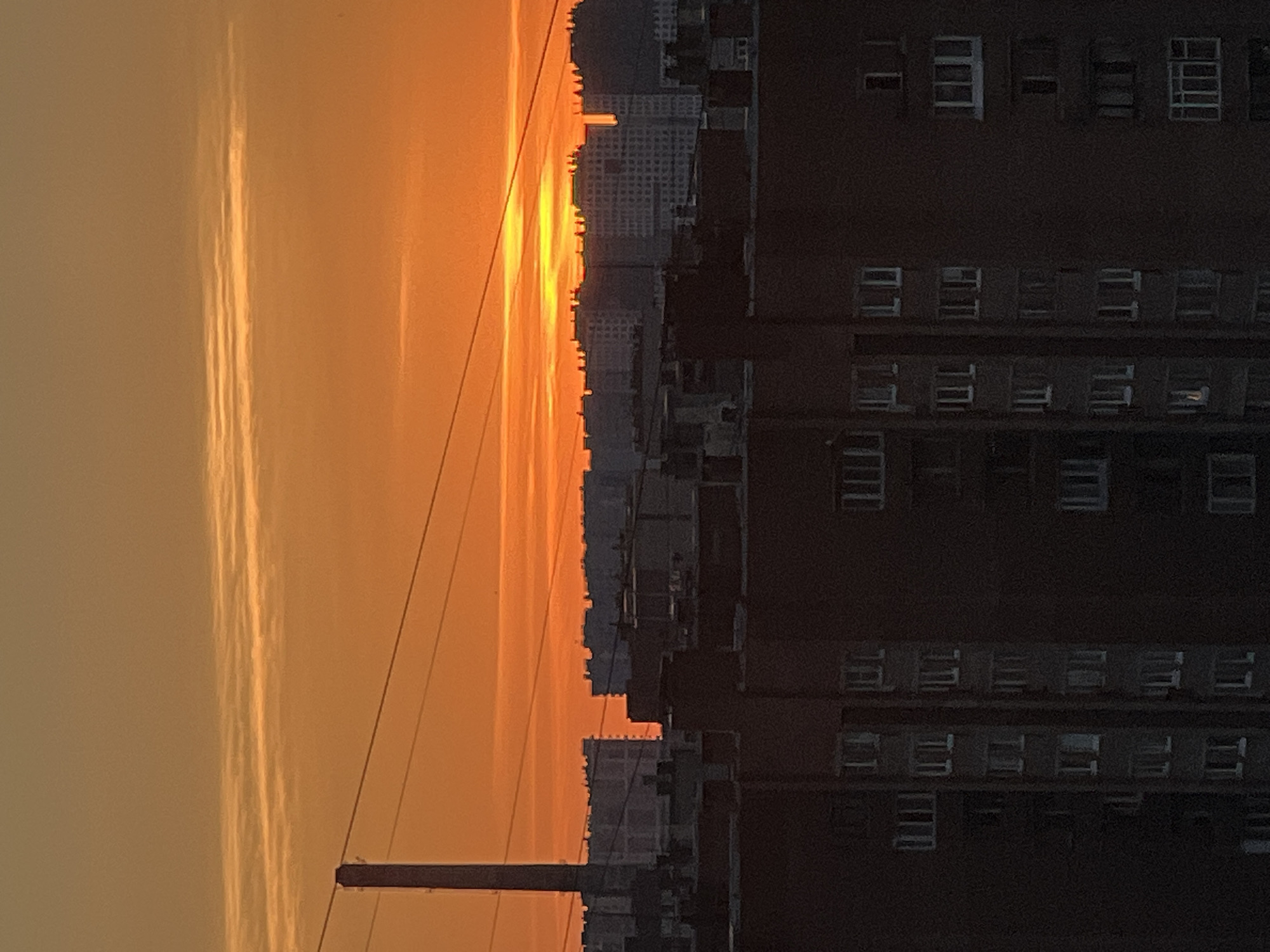 The image captures a sunset over the city of Sinyavino, Russia. In the foreground, silhouetted against the vibrant orange sky, are the dark forms of apartment buildings. These buildings are characterized by repetitive window patterns and are densely packed. A tall, slender chimney or smokestack is visible on the left side of the frame, also silhouetted. Several power lines stretch horizontally across the sky, adding a linear element to the composition. The sky itself is a gradient of orange and yellow hues, with thin, wispy clouds contributing to the atmospheric effect of the sunset. In the background, more apartment buildings of varying heights and architectural styles are visible, some appearing lighter in color and more distant. The overall scene depicts an urban landscape during twilight, dominated by the warm colors of the setting sun and the imposing structures of residential buildings. There are no people or active interactions visible in the image.