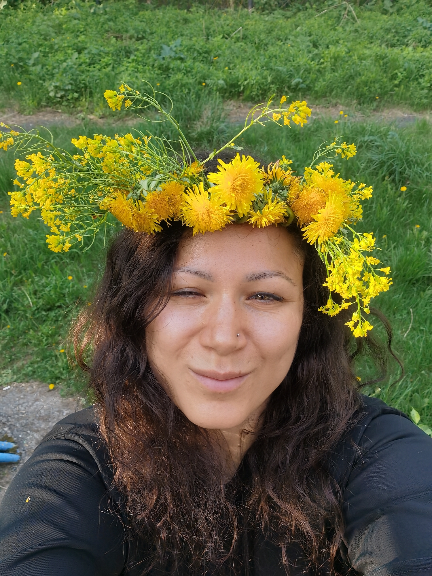 A woman with dark, curly hair is wearing a floral crown made of yellow dandelions and other yellow wildflowers. She is wearing a black shirt and has a nose piercing. Her right eye is closed, and she has a slight smile. The background is green grass with scattered yellow dandelions. There is a hint of a concrete surface and a blue object at the bottom left of the frame. The image appears to be taken outdoors in a park or grassy area, but there is no specific information to determine if it is in Perm, Russia.