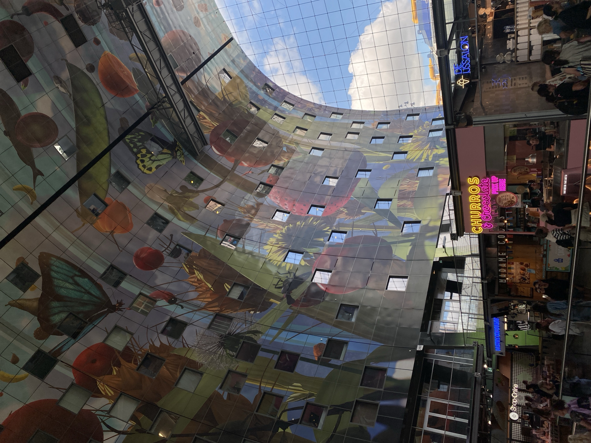 The image depicts the interior of the Markthal in Rotterdam, Netherlands. The ceiling features a large, colorful mural depicting fruits, vegetables, and butterflies, with numerous square windows interspersed throughout. Below the mural, a bustling marketplace is visible with various food stalls and shops. People are seen walking and standing around the market. One stall prominently displays a neon sign that reads "CHURROS & Chocolate". Another stall has a neon sign that says "DE JUSSALON". The architecture of the Markthal, with its curved structure, is evident. The overall scene is bright and lively, indicative of a public market.