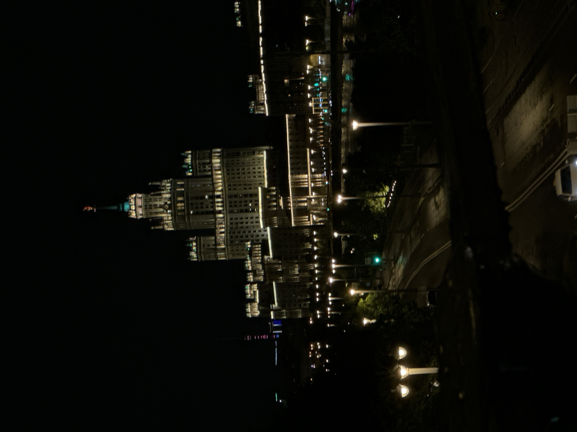 At night, a multi-story building dominates the left side of the frame. A road with cars lines the right of the frame, and street lights are placed between them. The building is identifiable as one of the Seven Sisters skyscrapers in Moscow, Russia.