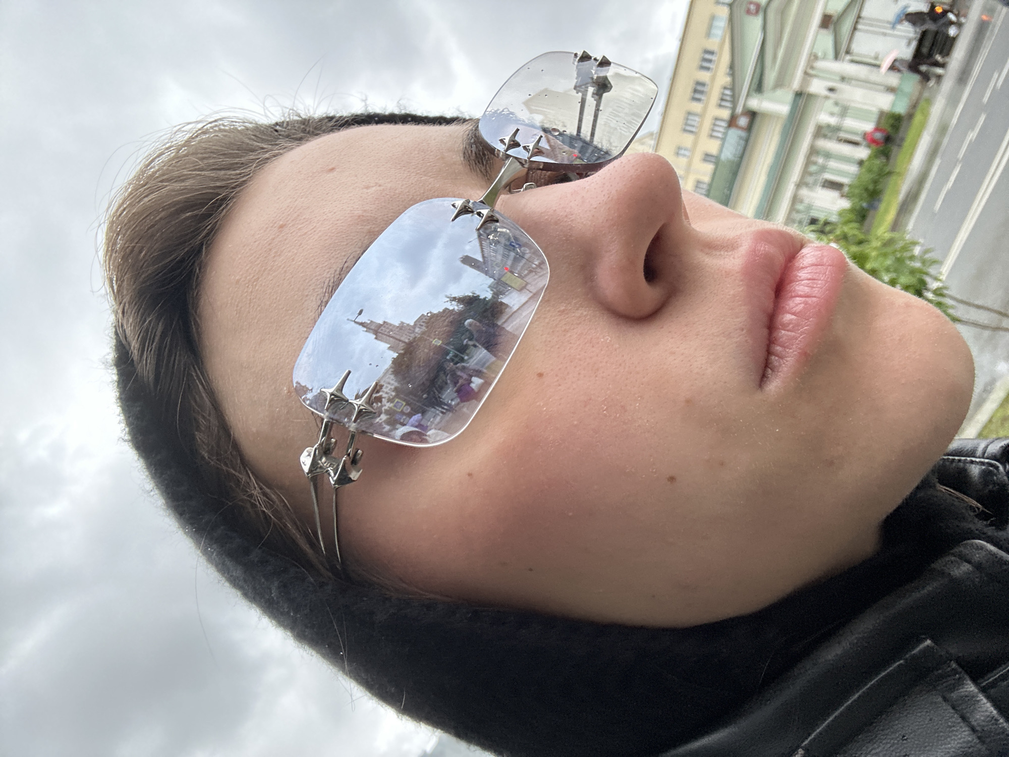 The image is a close-up, low-angle shot of a person wearing distinctive sunglasses with star-shaped accents on the frames. The person is wearing a black hooded jacket and a black leather jacket. The sunglasses' lenses reflect a street scene, including a tall, ornate building, people walking, and trees. The reflection suggests an urban environment with overcast skies. The background, though out of focus, shows a light-colored building with green architectural details, a street with white lane markings, and some greenery. Several people are visible in the background, some holding umbrellas, indicating it might be raining.
Based on the architectural style of the building reflected in the sunglasses and the general appearance of the surroundings, the scene is likely located in the vicinity of the Moscow State University (MSU) campus, possibly near one of its main buildings or a nearby historical structure. The ornate, classical architecture of the reflected building strongly resembles the Stalinist Empire style characteristic of Moscow's Seven Sisters skyscrapers, with MSU being the most prominent example. The presence of trees and a street layout also aligns with the areas surrounding MSU's main building.