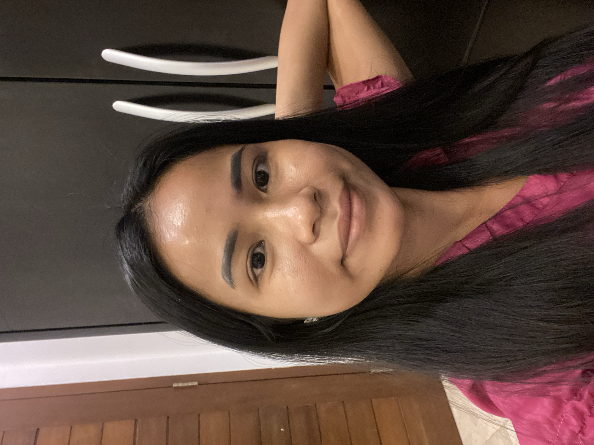 A woman with long, straight black hair is posing for a selfie. She is wearing a pink shirt. The background includes a dark gray cabinet with white handles, a white wall, and a light brown wooden door. The photo was taken indoors. The location may be North Kuta, Indonesia, based on user input.
