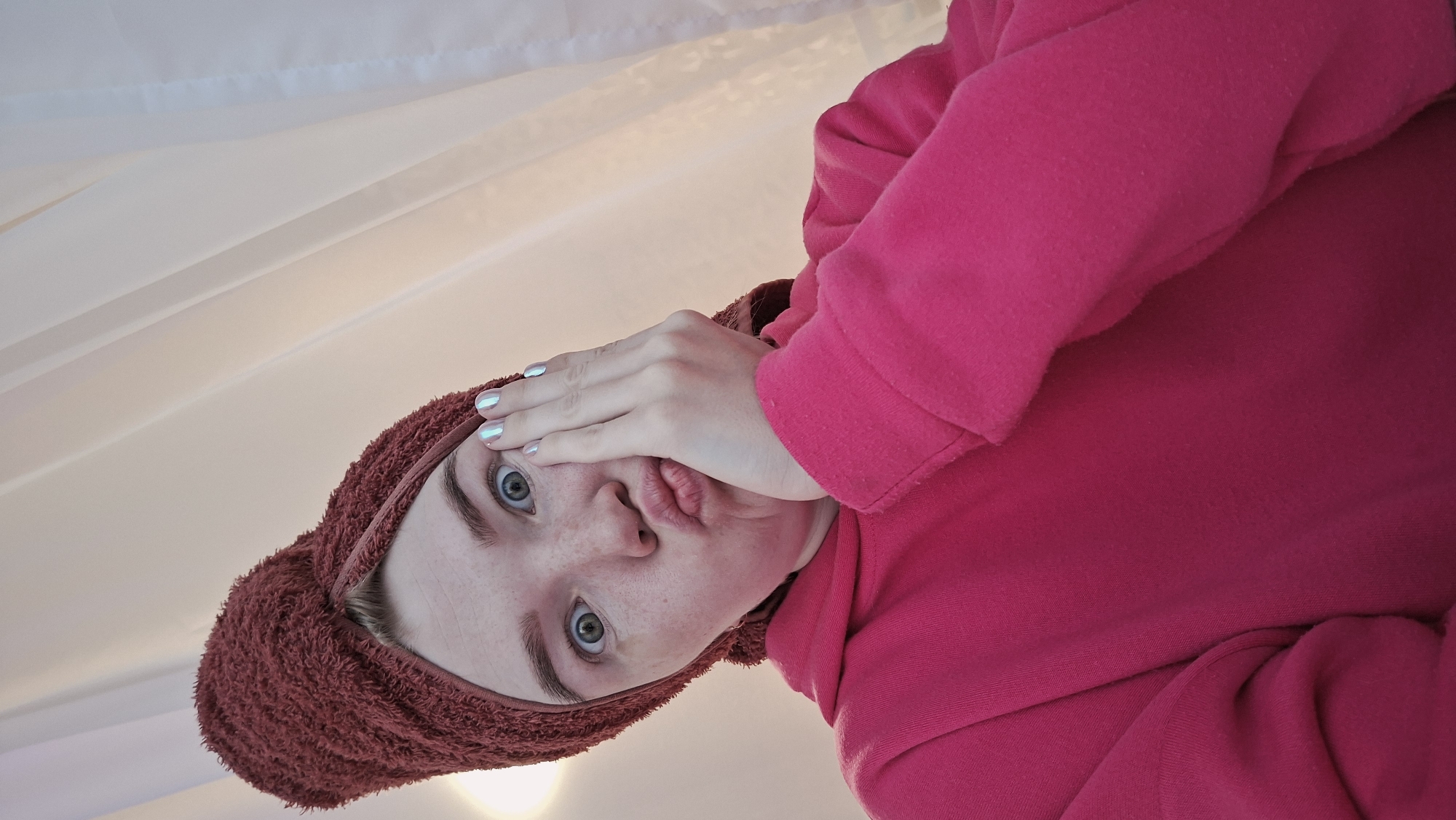 A young person with fair skin, wearing a red hooded sweatshirt and a dark red towel wrapped around their head, is posing for a close-up photograph. The person is looking directly at the camera with wide eyes and pursed lips, covering part of their face with their hand. The nails are painted with a light, iridescent color. The background appears to be a white surface with subtle shadows. The scene is likely indoors. There is no verifiable information to confirm that the image was taken in Chaikovsky, Russia.