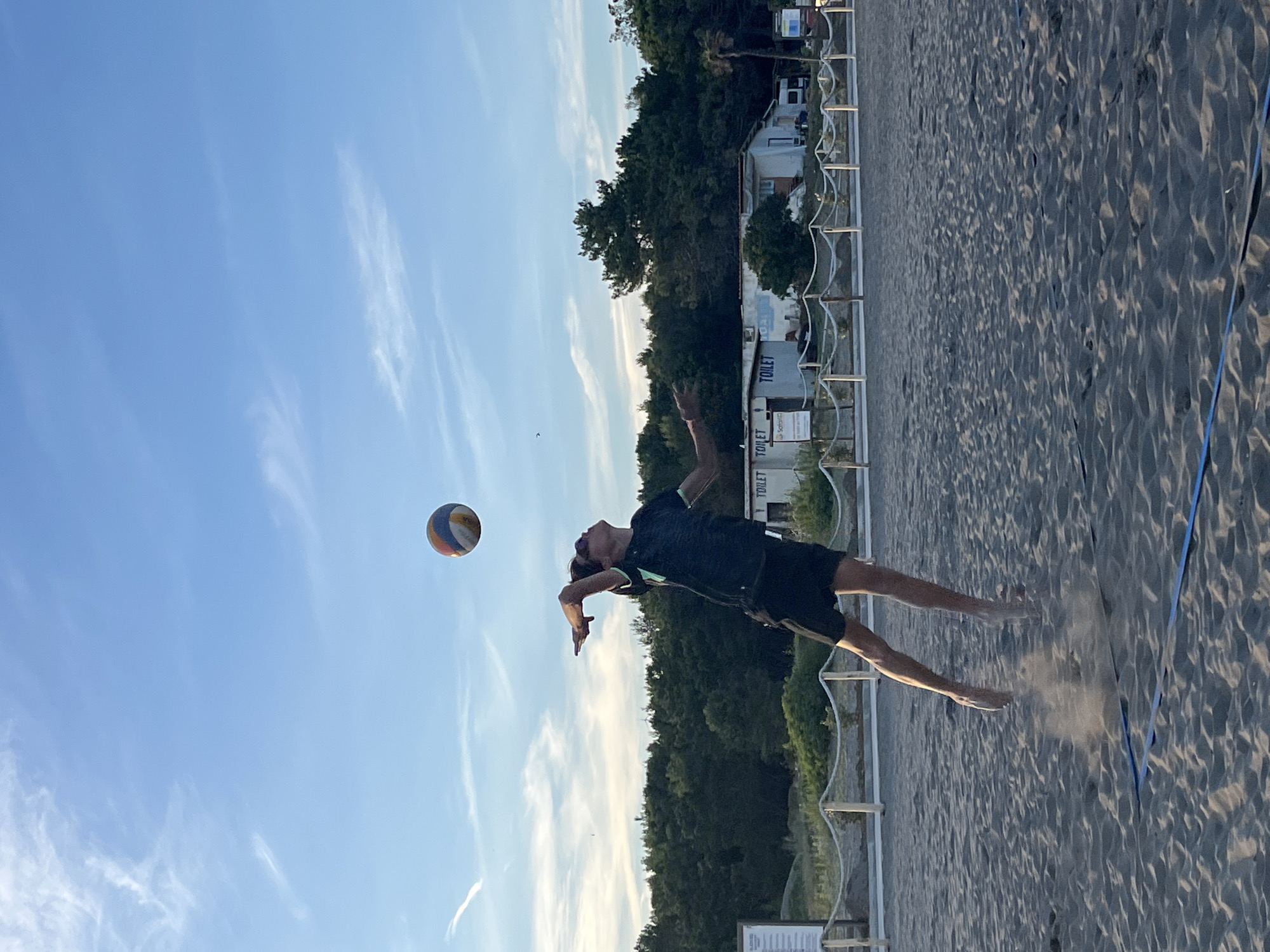 A young person is captured mid-air on a sandy beach court, executing a volleyball serve. The player, wearing a dark t-shirt with a lighter trim and black shorts, is focused on the ball suspended above and slightly in front of them. Their left arm is extended upwards, and their right arm is brought back, poised to strike. The sand is kicked up around their feet, indicating the power of their jump. In the background, a line of trees forms a dense green backdrop against a clear blue sky with wispy white clouds. Several small, white buildings with "TOILET" signs are visible behind a rope fence that separates the court from the background area. The beach volleyball court has visible blue boundary lines on the sand. The scene appears to be set in the city of Pistula, Montenegro, during daylight.