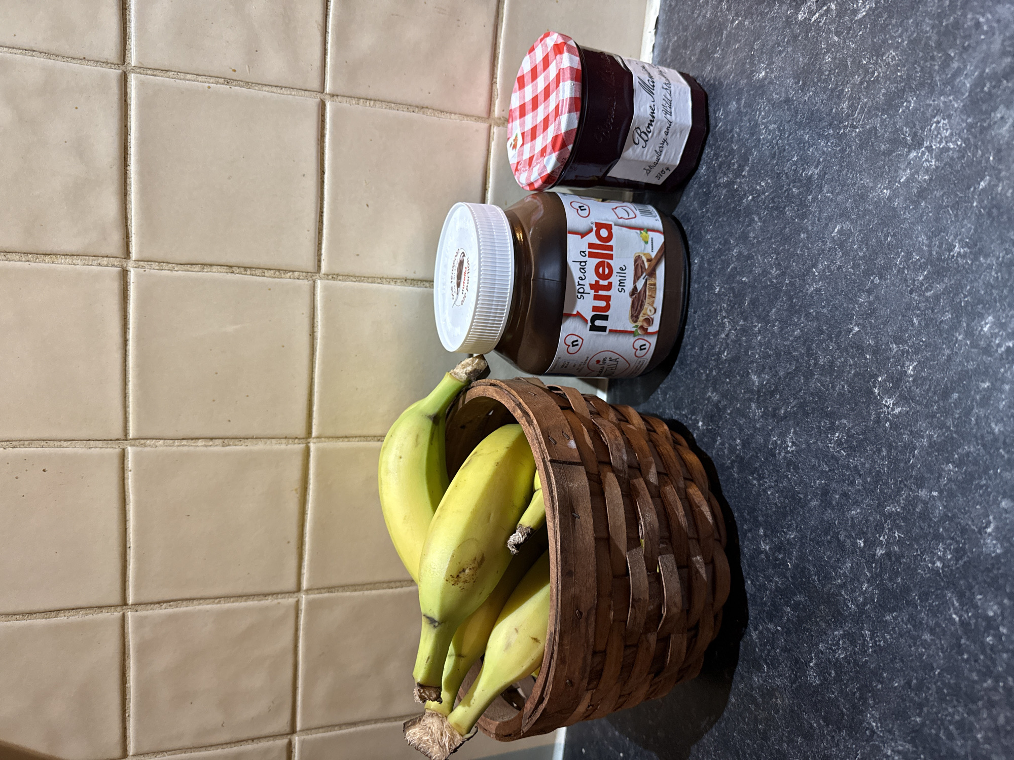 The image shows a kitchen scene. A woven basket containing several unripe bananas sits on a gray countertop. Above the basket are two jars. The lower jar is labeled "Nutella," and the upper jar has a red and white checkered lid. The background behind the basket and jars is tiled with beige square tiles. The location of the image is not visible in the picture, so I cannot confirm it is Great Yarmouth, United Kingdom.