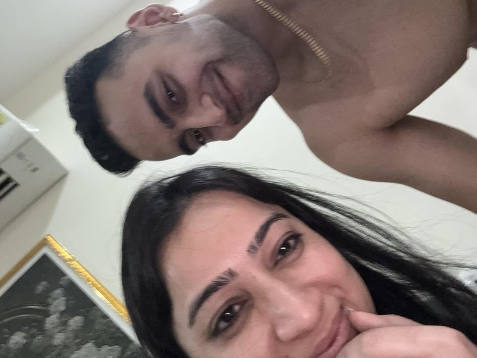The image depicts two individuals in what appears to be an indoor setting in Malasar, India. In the foreground, positioned to the left, is an adult female with dark hair and dark eyes, looking directly at the camera with a slight smile. Her right hand is positioned near her chin/mouth. To her right and slightly behind her, an adult male with short dark hair, dark eyes, and light facial hair is visible, also smiling and looking towards the camera. He is shirtless, displaying his chest and right shoulder, and wears a gold-colored chain around his neck and a small gold hoop earring in his left ear. The background consists of a light-colored wall. Partially visible in the upper left corner is a gold-framed artwork, and an off-white air conditioning unit is mounted high on the wall in the upper center-right.