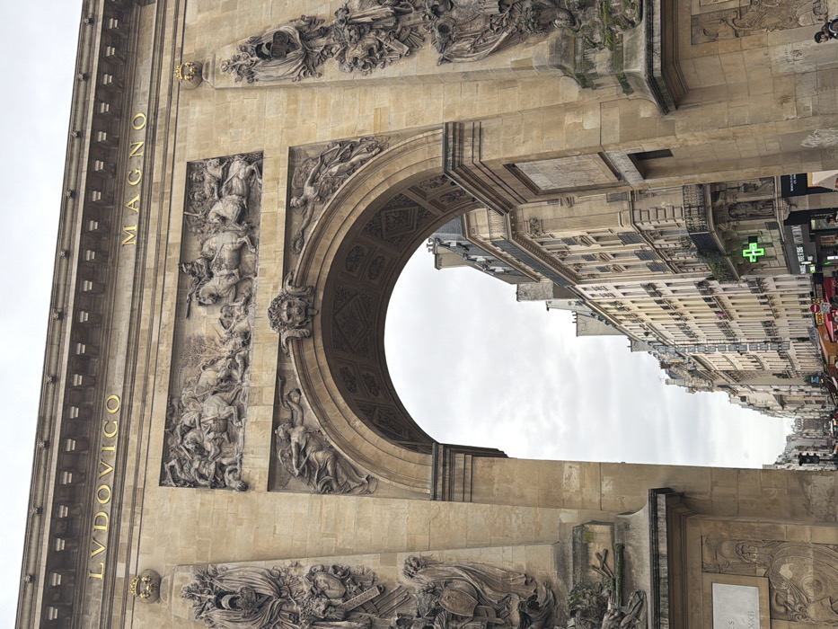 The image displays a close-up, slightly upward-angled view of the Porte Saint-Denis, a triumphal arch situated on the Rue du Faubourg Saint-Denis in Paris, France. The arch is constructed of light-colored stone, exhibiting intricate classical architectural details. Above its large central archway, a horizontal frieze features a high-relief carving depicting a dynamic battle scene with multiple mounted and dismounted figures engaged in combat. Above this frieze, a prominent cornice supports a horizontal band with gilded capital letters spelling "LVDOVICO MAGNO." The keystone of the archway is adorned with a carved lion's head, flanked by two relief sculptures of draped, winged figures. The arch's side piers contain vertical relief panels depicting military trophies, shields, and drapery.
Through the archway, a perspective of a Parisian street is visible under an overcast, gray sky. The street is lined with multi-story, light-colored buildings of typical Haussmannian architectural style, featuring numerous windows and small balconies. On the right side of the street, at ground level, a bright green illuminated cross, characteristic of a pharmacy, is clearly visible. Distant street-level activity and what appear to be awnings or market stalls are present. No distinct individuals or specific actions are discernible in the immediate foreground or midground; distant figures on the street are indistinct.