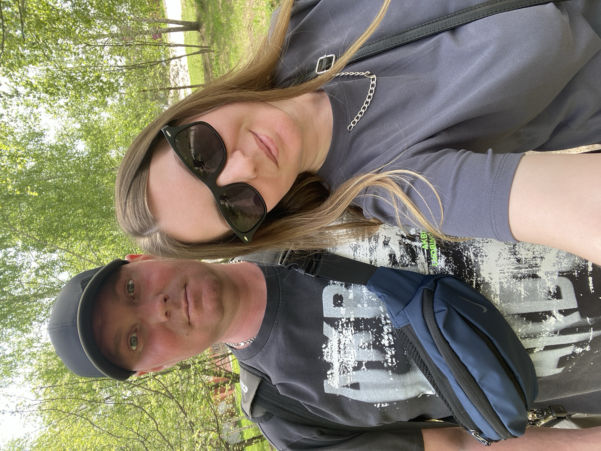 Two individuals are captured in an outdoor setting, presumably in Noviy, Russia. The woman is positioned above the man, possibly in a selfie pose, wearing dark sunglasses and a gray top adorned with a chain detail. The man is below, wearing a dark baseball cap and a printed T-shirt with a blue Nike fanny pack across his chest. The background suggests a park or green space with trees and foliage.