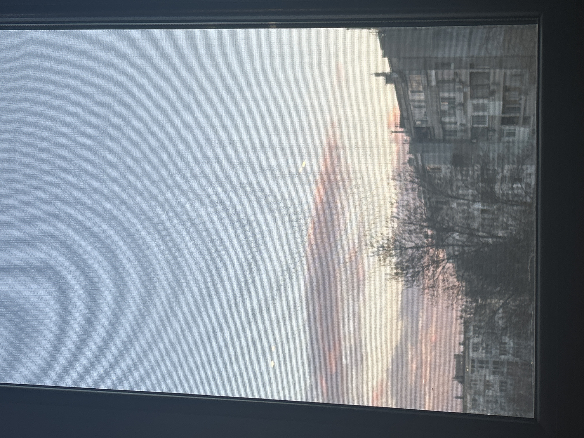 The image, taken through a window screen, depicts an urban landscape. The sky is light blue, transitioning to pink and orange hues near the horizon, indicating sunrise or sunset. Visible through the screen are apartment buildings, characteristic of urban residential architecture. Silhouetted tree branches are visible in the foreground, framing the buildings. The scene is likely located in Varna, Bulgaria, based on the architectural style of the apartment buildings. The window frame is dark, contrasting with the bright outdoor scene. No people are directly visible in the image. The interaction captured is between the natural light and the built environment, viewed through the filter of the window screen.
