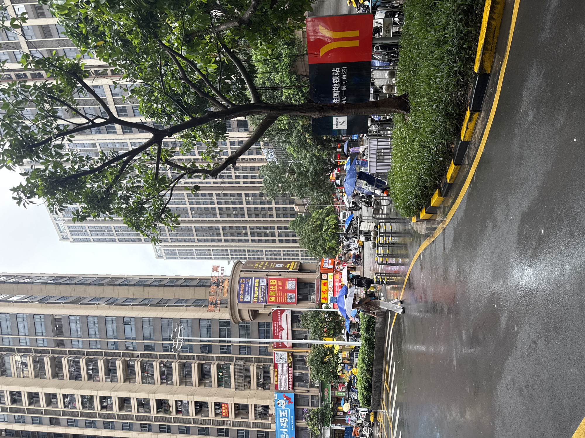 The image depicts a street scene in Haizhu District, China, during a light rain. The foreground features a wet asphalt road with yellow and black painted curbs, indicating a street corner with a pedestrian crossing. Several parked electric scooters and motorcycles are visible on the right side of the road. In the midground, a group of people are walking, some holding umbrellas. A woman in white pants and a brown top is walking across the pedestrian crossing, holding a black umbrella. A large tree with green foliage stands prominently on the right side of the frame, partially obscuring the background. Behind the tree, a black and red sign with Chinese characters and a yellow arrow pointing left is displayed. The background is dominated by tall, modern apartment buildings with numerous windows and balconies. Several smaller buildings with commercial signage are also visible. The sky is overcast and cloudy, consistent with rainy weather. The overall impression is of a bustling urban environment experiencing a day of rain.