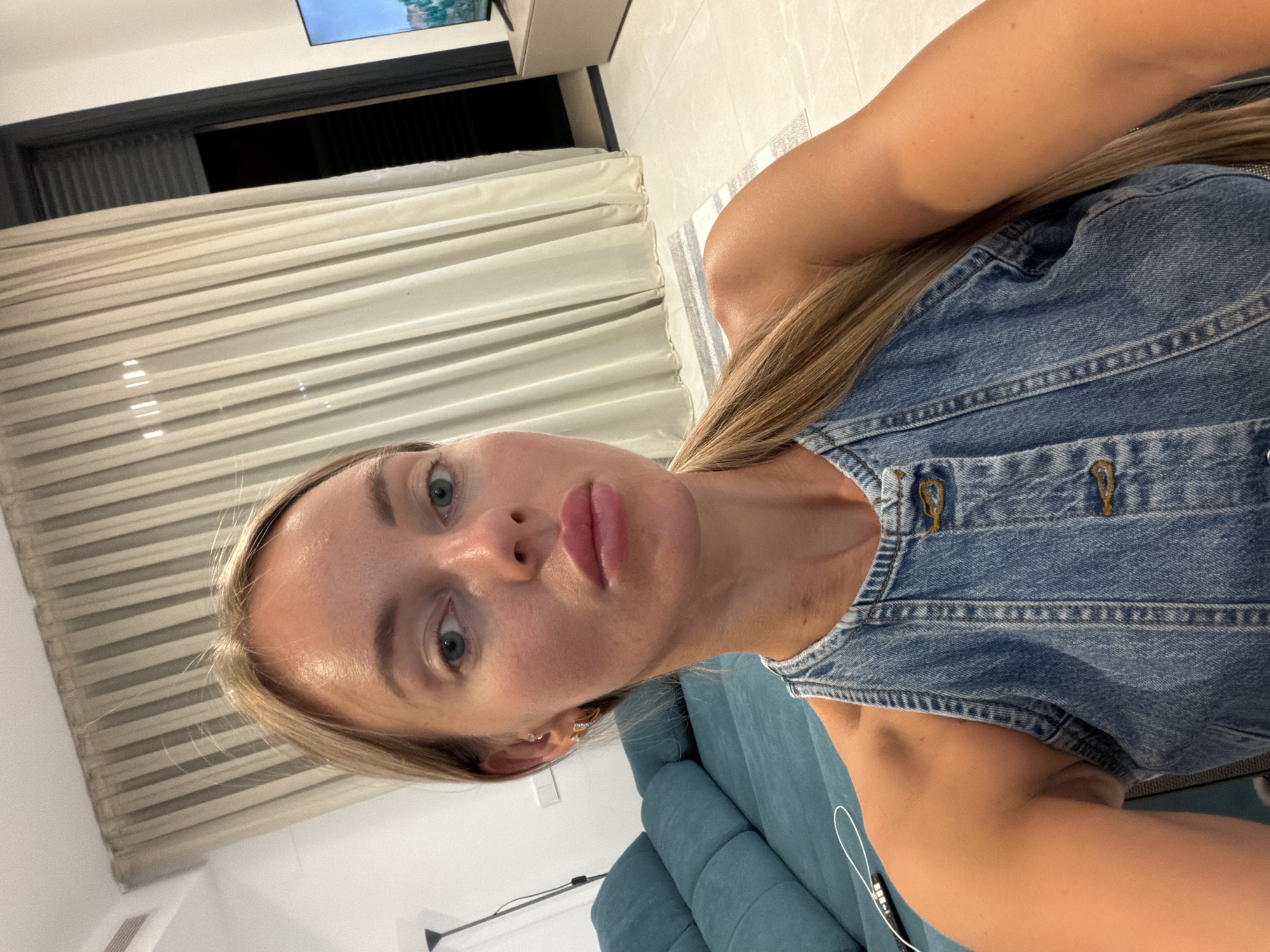 A woman with blonde hair pulled into a ponytail, wearing a denim top, is pictured from the chest up in a selfie. She is looking directly at the camera. Behind her, a teal sofa is visible, along with sheer beige curtains covering a window. The scene appears to be indoors, possibly in a living room, in Finestrat, Spain.