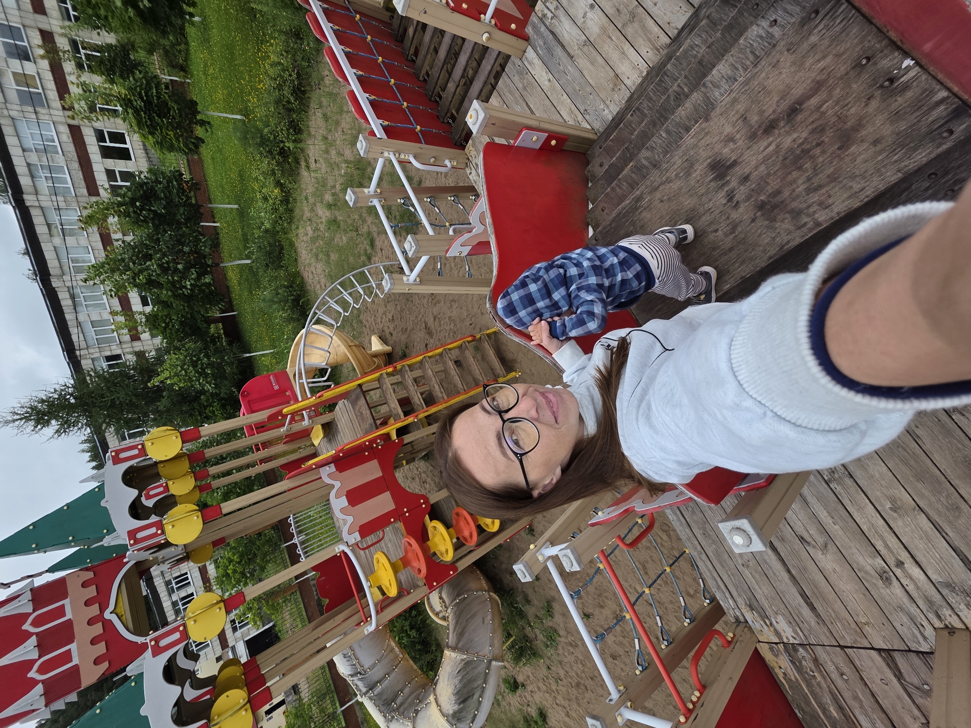 In Chaikovsky, Russia, a woman with glasses and a young child in a plaid shirt are at a playground. The playground features a large, colorful castle-themed structure with slides, bridges, and climbing elements. The woman is taking a selfie while holding the child's hand. They are on a wooden platform of the playground. The background shows a school building and green trees. The weather appears overcast.