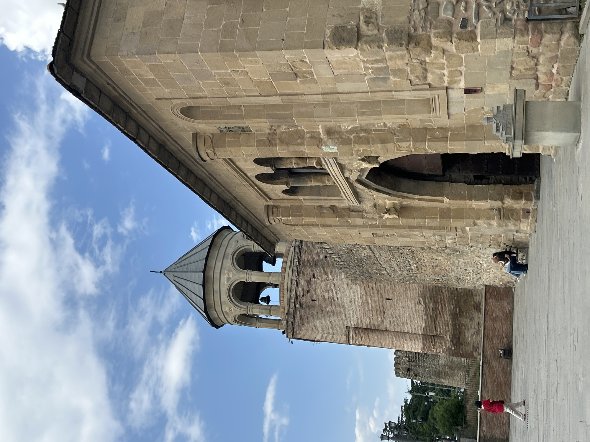 This is a medium-angle, eye-level shot of the Svetitskhoveli Cathedral complex in Mtskheta, Georgia, on a partly cloudy day. The foreground is a wide, paved courtyard. To the right is the stone wall of the cathedral, with a large arched entryway visible. Above the entryway are arched windows with decorative columns. To the left is a tall, brick bell tower topped with a conical roof and a series of arched openings. Two bells are visible within the tower's belfry. In the courtyard, a person in a red top and cream pants walks from left to right. Further back, two people are seated on a bench to the right, with one person appearing to gesture. A small model of a cathedral sits on a pedestal in front of the stone wall. Trees and a section of a fortified wall are visible in the background.