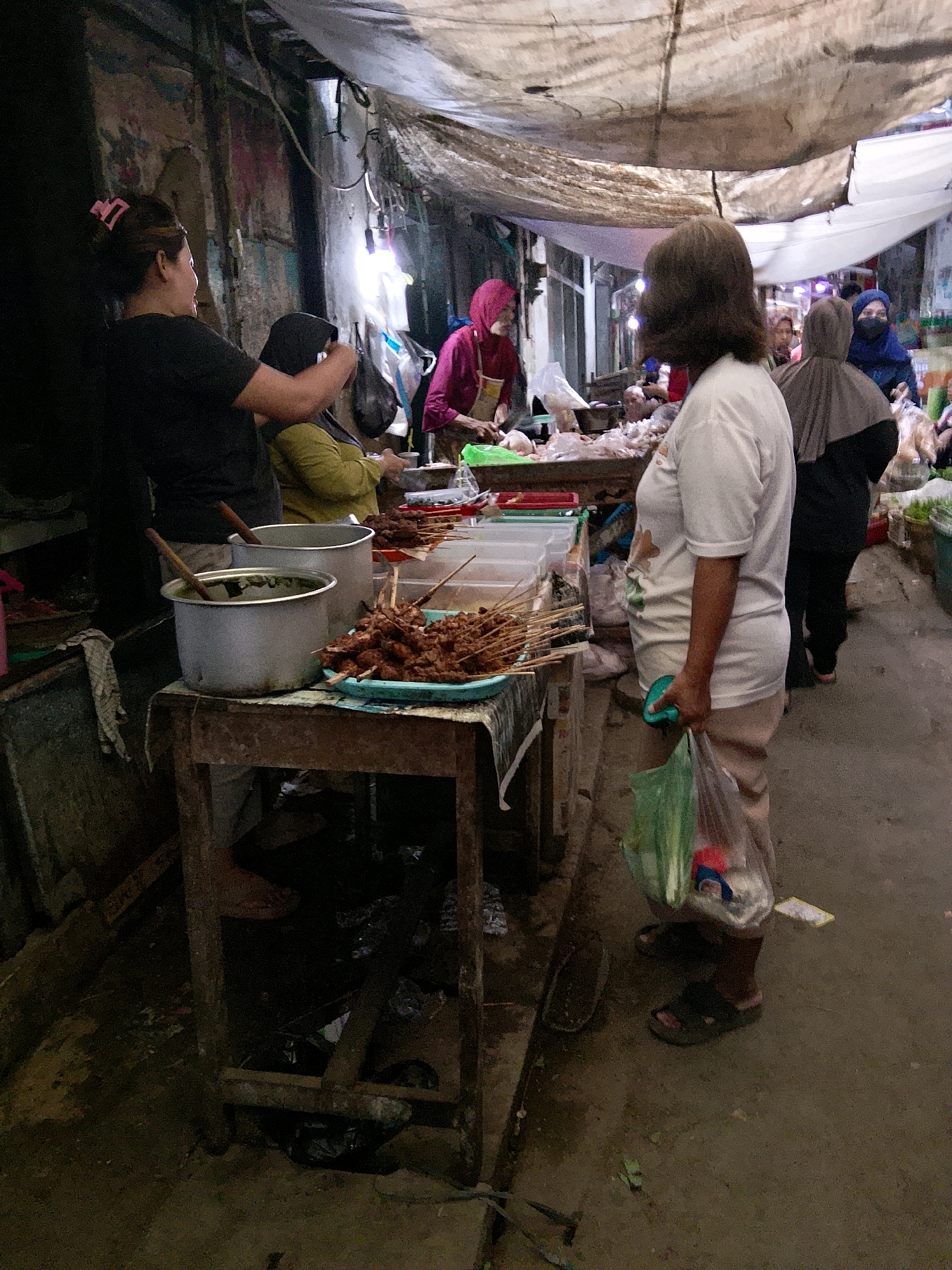 A vendor stands behind a stall in a market in Cirebon, Indonesia, preparing food. She is wearing a white t-shirt and light-colored pants, holding a green plastic bag in her right hand. The stall is covered with a tarp, and a variety of food items are displayed, including skewers of meat. Several other people are visible in the background, some wearing hijabs. The ground is paved, and the overall impression is one of a busy, outdoor market.