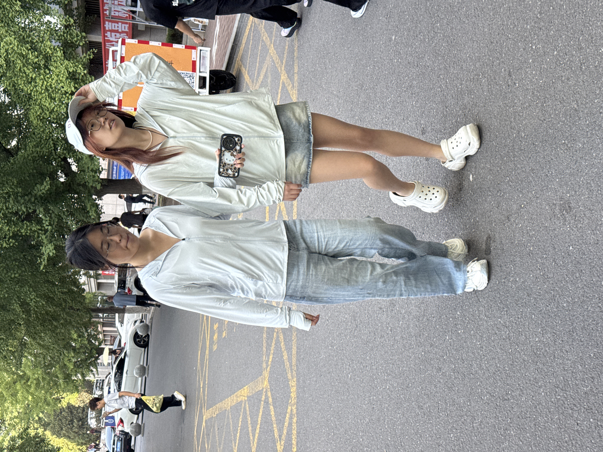 Two young women are walking on a paved street in what appears to be the city of Zhongtun, China. The woman on the left is wearing a light blue zip-up hoodie, light blue wide-leg jeans, and white sneakers. She has dark hair pulled back and wears glasses. The woman on the right is wearing a similar light blue zip-up hoodie over a denim skirt and white platform clogs. She has long reddish-brown hair, wears glasses, and a white baseball cap. She is holding a smartphone in her right hand. The street is marked with yellow lines, and trees line the background. Cars and buildings are visible in the distance. The lighting suggests it is daytime.