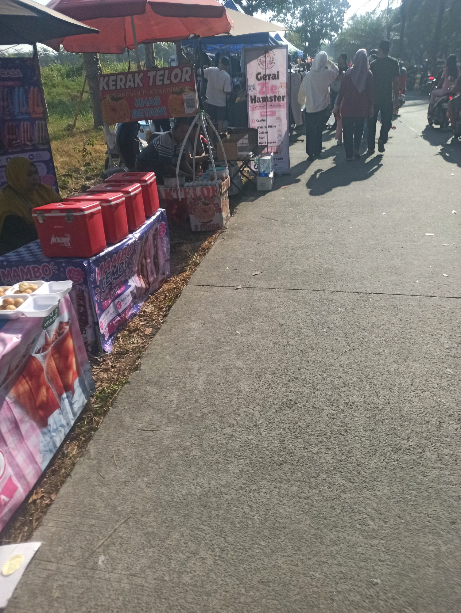 This is a medium-shot, eye-level, vertical, outdoor photograph of a street market or roadside vendors. The scene is set in Telagamurni, Indonesia.
In the foreground, on the left, is a stall with several red coolers on top of a table covered with a blue and white checkered cloth. A person wearing a yellow hijab is partially visible behind the coolers, looking to the left. In front of this stall, another table is set up with white styrofoam containers filled with small, golden-brown, round food items. A portion of a pink and white patterned cloth covers the front of this table, with an image of a drink printed on it.
Moving towards the center of the image, a stall is set up with a sign that reads "KERAK TELOR MAKANAN TRADISIONAL KHAS BETAWI." Next to this is another stall with a vertical banner advertising "Gerai Zie Hamster." Several people are walking past these stalls. One person in the foreground is wearing a white hooded jacket, another in a maroon hijab and jacket, and a third person in a dark t-shirt. Shadows of these individuals are cast on the asphalt road.
Further in the background, more people are visible walking and standing. Several motorcycles are parked on the right side of the road. The environment suggests a sunny day with trees lining the background. The road is paved with asphalt and shows signs of wear. The overall impression is a bustling local market with various food and merchandise stalls.