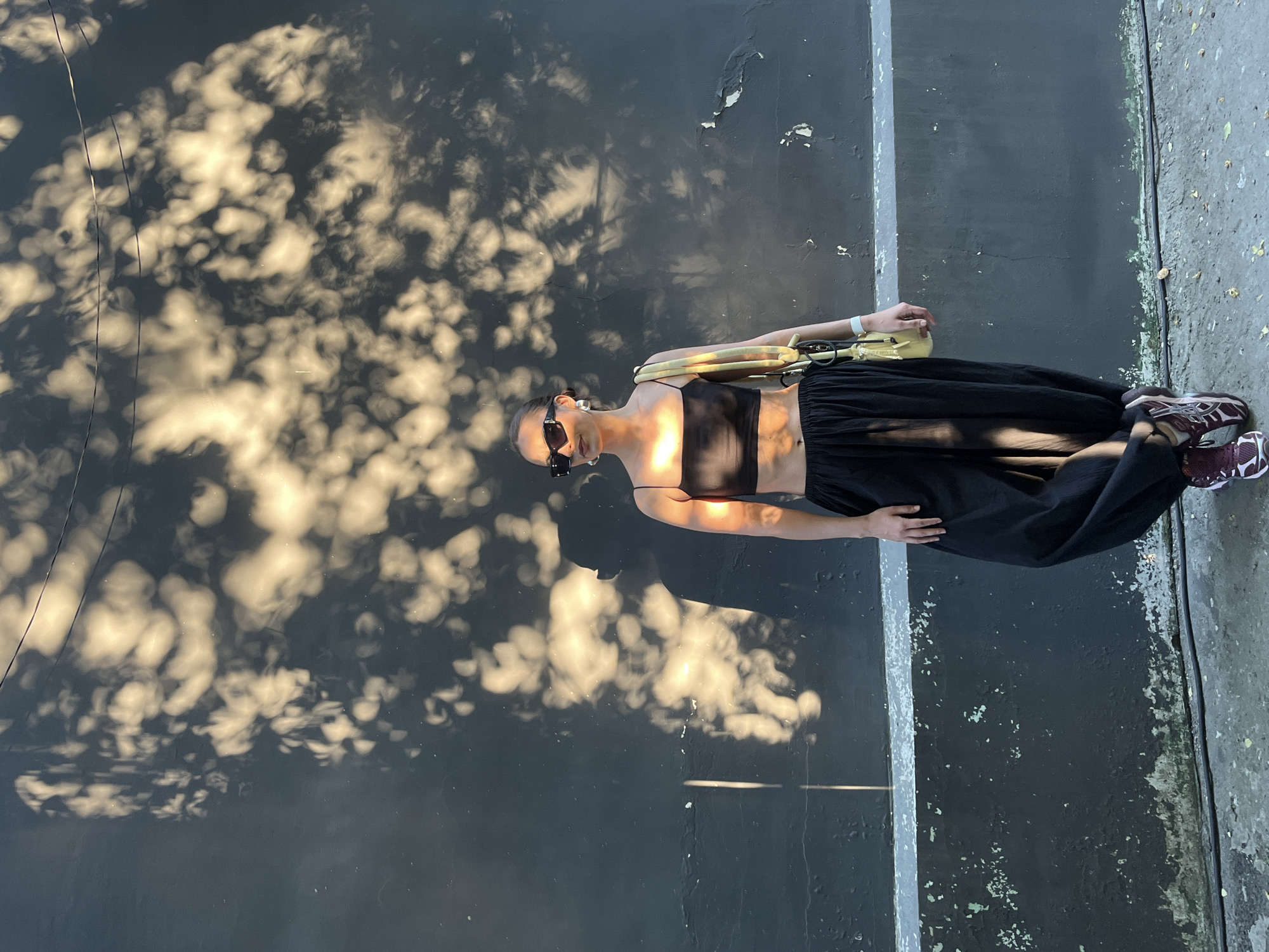 The image depicts a full-body shot of an individual, likely a woman, standing outdoors against a dark grey or black textured wall. The person is positioned centrally, facing slightly left with their head turned forward, obscured by large rectangular black sunglasses. They have dark hair pulled back and wear large silver hoop earrings. The individual is dressed in a black, thin-strap crop top and loose-fitting black wide-leg trousers. On their left wrist is a white band or watch, and a light yellow/beige hobo-style shoulder bag with a thick, textured strap hangs over their left shoulder. They are wearing burgundy and white athletic sneakers.
The environment is characterized by a dark wall, which shows signs of wear, including peeling paint or plaster, particularly along its lower edge, and some green/white discoloration where it meets the ground. A distinct horizontal white or light grey stripe runs across the wall at mid-height. The ground beneath is concrete or asphalt, with scattered debris or small leaves, and a dark wire or cable runs along its base. The scene is illuminated by strong, warm sunlight, casting prominent, dappled shadows of tree leaves or foliage across the entire wall and parts of the person's body. Bright spots of direct sunlight penetrate the leaf canopy, creating luminous patterns. A large shadow of a person, presumably the subject, is cast on the wall to their immediate left. In the upper portion of the frame, two thin, dark overhead wires are visible against the wall. The specific city of Krasnoyarsk, Russia, cannot be determined from the visual information provided in the image.
