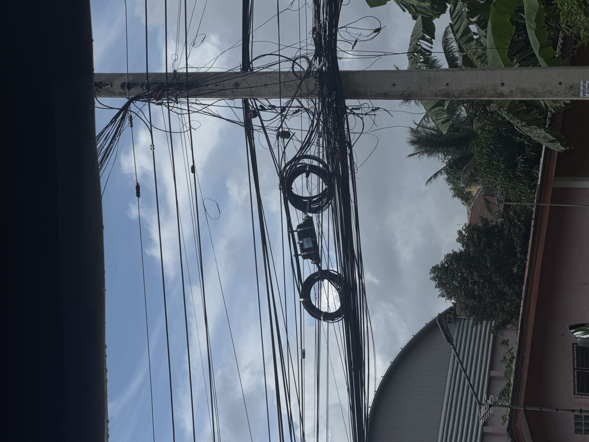 The image captures a dense network of overhead power and telecommunications cables strung between concrete utility poles against a cloudy sky in Kathu, Thailand. The cables are numerous, tangled, and organized haphazardly, characteristic of urban infrastructure in the region. A black box, likely a junction or splitter for fiber optics, is visible, suspended among the wires. In the foreground, the roofline of a building with a corrugated metal structure and a pink facade is partially visible, along with lush green foliage, including palm leaves and trees, suggesting a tropical environment. A street lamp and antenna are also present.