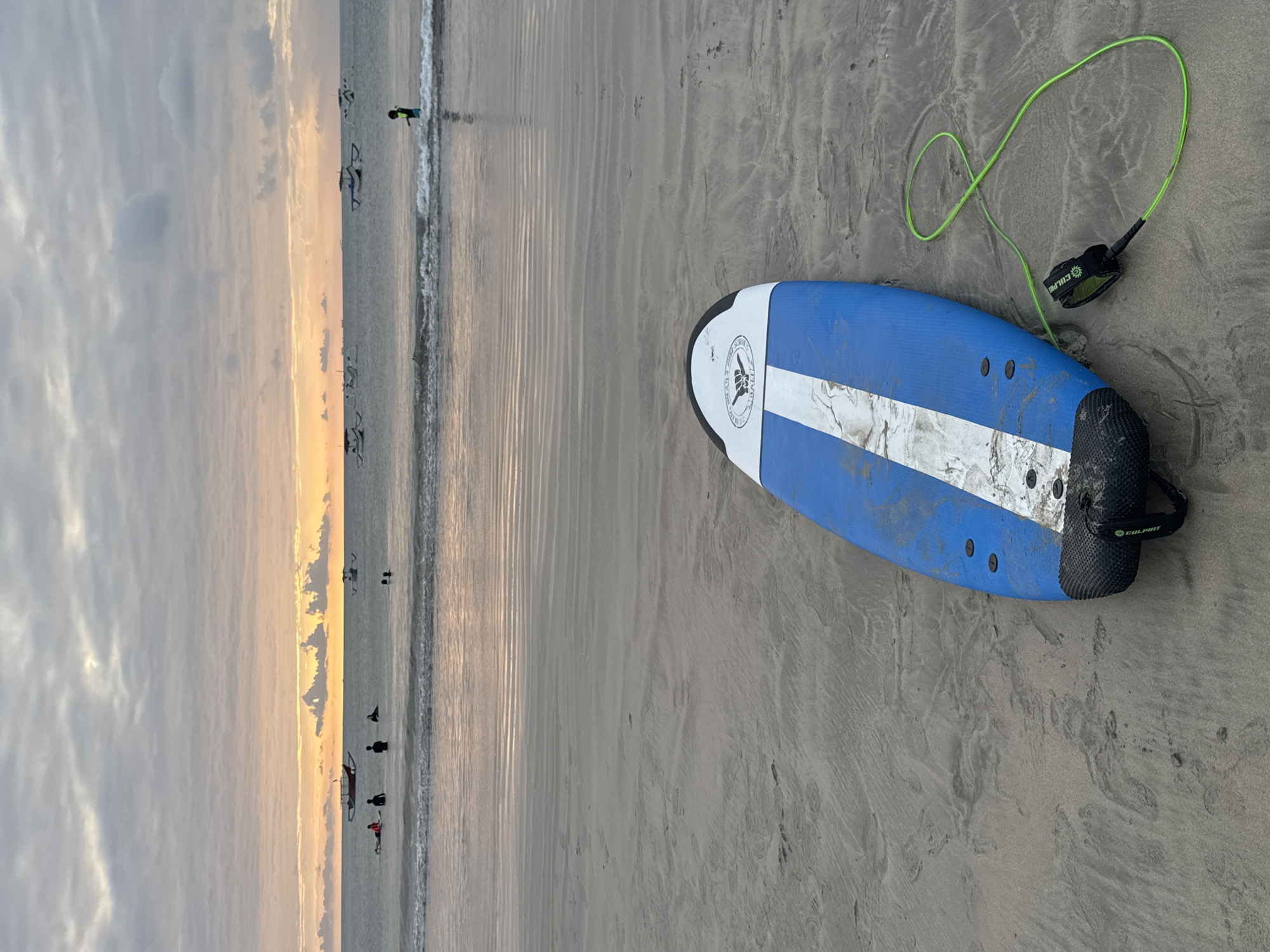 A blue and white surfboard with a green leash lies on a sandy beach in Kuta Selatan, Indonesia, during a sunset. In the background, several traditional fishing boats are visible on the ocean. A few people are in the water, some of whom appear to be surfing. The sky is cloudy with a warm orange glow from the setting sun. The tide is low, leaving a wet, reflective surface on the sand.