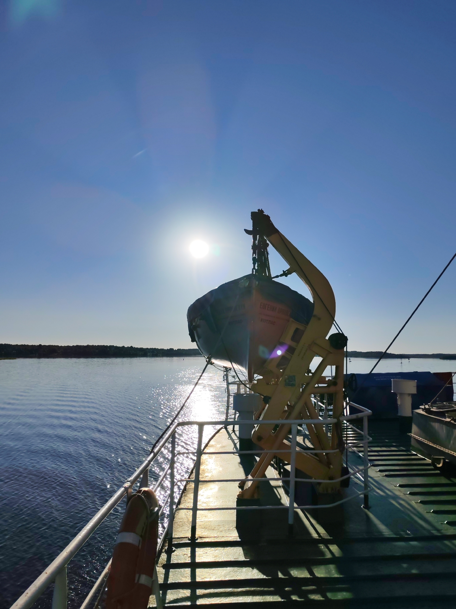 The image depicts a bright, sunny daytime scene observed from the deck of a vessel navigating a calm body of water, presumably in the vicinity of Mys, Russia. The sky is clear, deep blue, with the sun prominently visible in the upper right quadrant, emitting strong glare and lens flares. A faint, thin white contrail is discernible in the upper-central sky.
The body of water, likely a river or large lake, displays gentle ripples and intense specular reflections from the sun, creating a shimmering effect, particularly on the left side and extending towards the distant horizon. A dark, densely treed shoreline or landmass is visible on the distant horizon, primarily on the left side of the frame.
In the foreground, the dark green deck of the vessel is visible, featuring parallel textural lines. A white/silver metal railing runs along the port side from the lower left to the middle right. An orange lifebuoy with white reflective tape hangs from this railing in the lower-left quadrant.
A dark blue/grey enclosed lifeboat or rescue capsule is suspended from a large, prominent yellow davit system, positioned mid-deck. The lifeboat features Cyrillic inscriptions: "ЕВГЕНИЙ ДРОНОВ" (Yevgeniy Dronov), likely the name of the vessel or lifeboat, and "КОТТЛАС" (KOTTLAS), indicating a homeport or registry.
Behind the davit, covered objects under blue tarpaulins are visible on the deck, alongside a white vertical vent pipe. Dark diagonal lines, likely rigging or mooring ropes, descend from the upper right corner towards the deck. The sun's position casts long, distinct shadows from the railing, davit, and other deck structures across the deck towards the left. No individuals are visible in the frame.