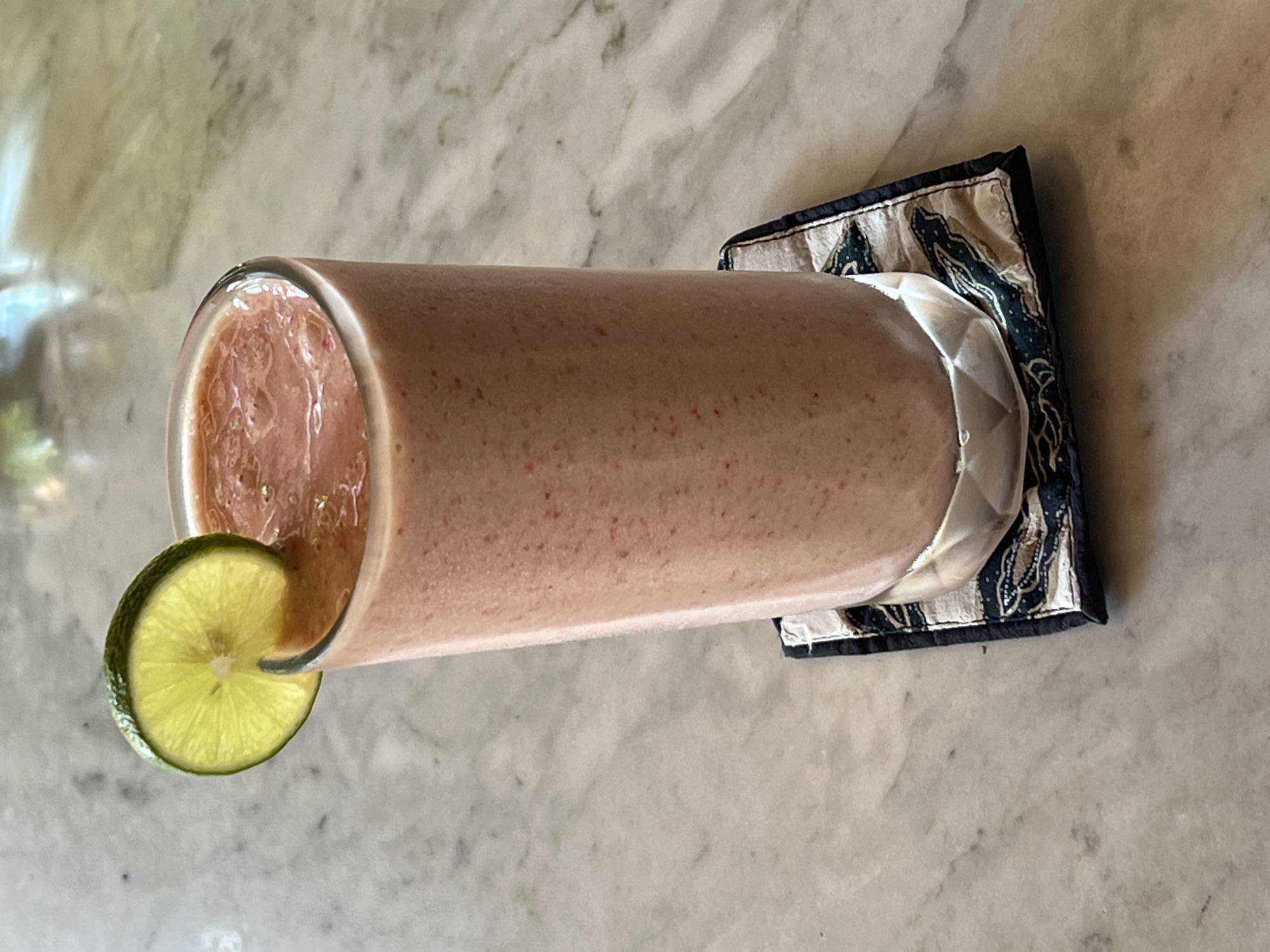 A tall glass of pink strawberry smoothie sits on a patterned coaster on a marble countertop. A slice of lime is perched on the rim of the glass. The background is blurred and suggests an indoor setting, possibly a cafe or restaurant in Ubud, Indonesia.