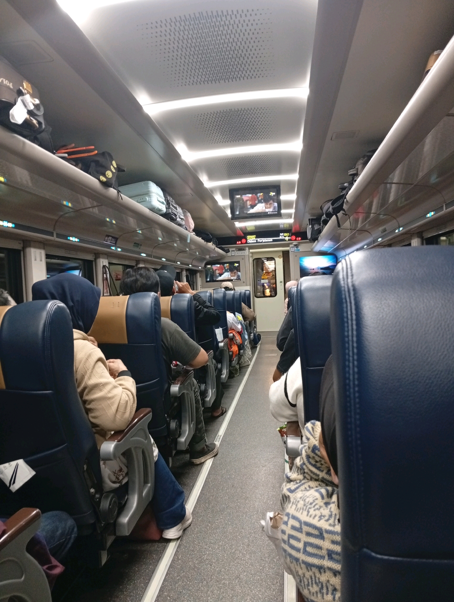 The image shows the interior of a train carriage with passengers seated on blue and tan seats. Luggage is stored on overhead racks. A screen mounted on the ceiling displays an image of a person. The lighting appears to be artificial, with recessed lights along the ceiling. The train appears to be moving, as suggested by the blur in some parts of the image, however, this is not definitive. The overall atmosphere is that of travel in Indonesia.