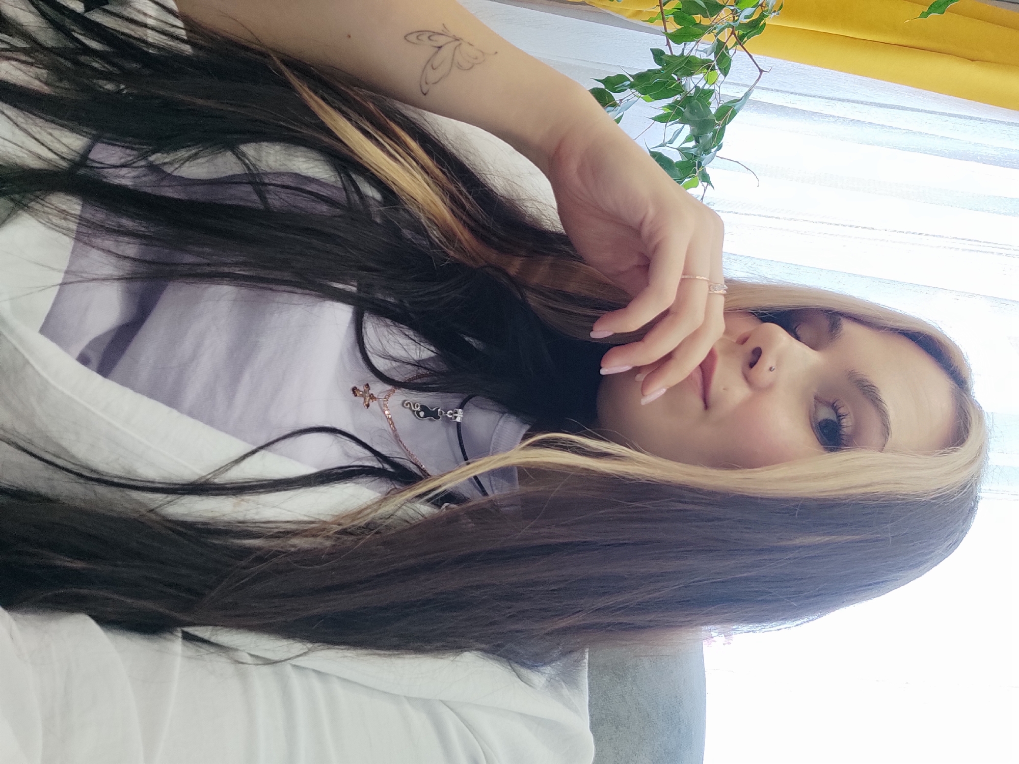 A young woman with black and blonde-streaked hair is looking at the camera with a serene expression. She is wearing a light-colored t-shirt and has a butterfly tattoo on her arm. She is also wearing a nose ring and a ring on her finger. There are some plants and a yellow curtain visible in the background. The location is not definitively identifiable as Ольховка, Russia based on the provided image.