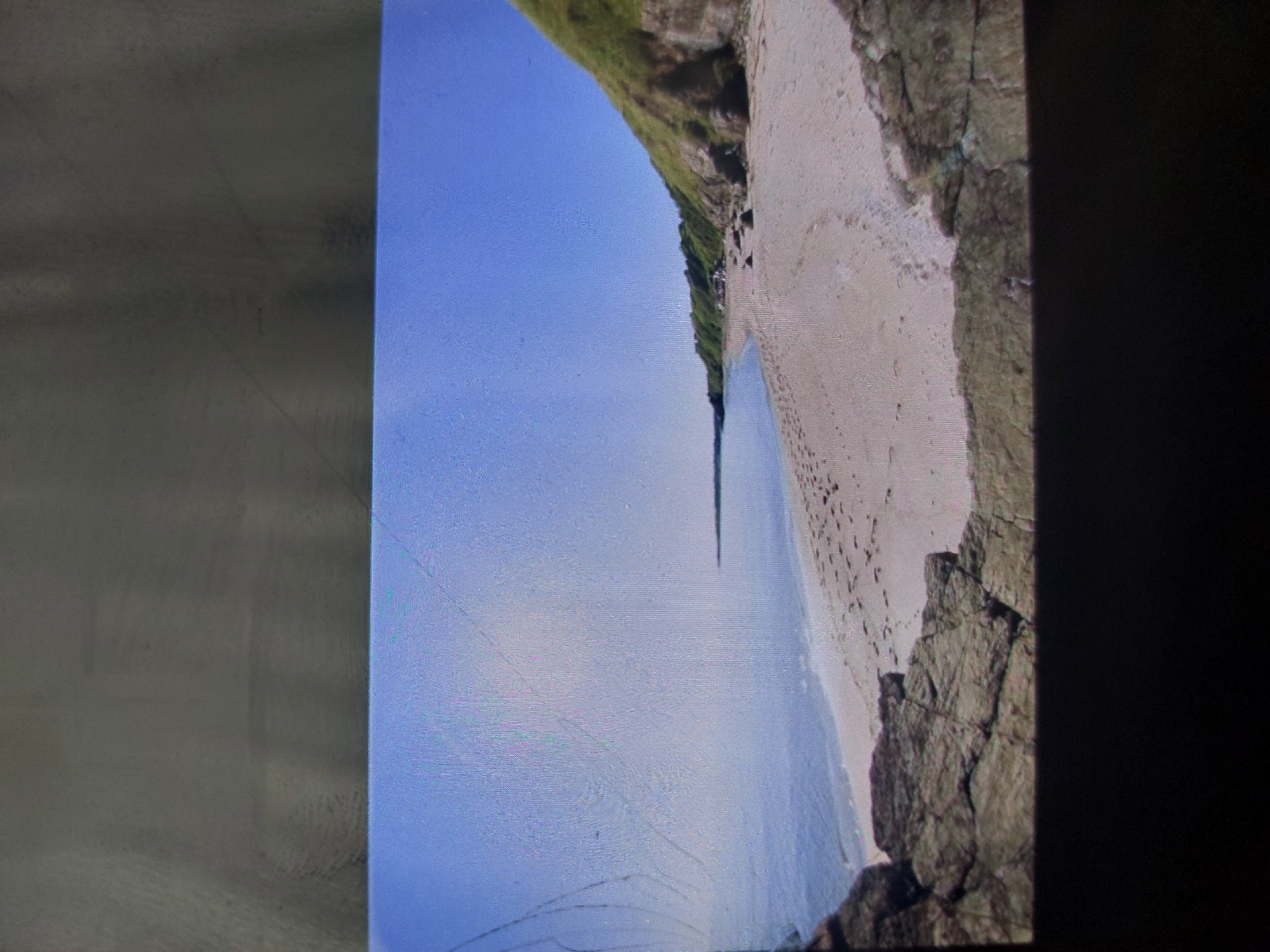 The image depicts a beach scene. A sandy beach stretches along the coastline, bordered by rocky cliffs and greenery on one side, and the blue sea and sky on the other. Footprints are visible on the sand. The specific location is not discernible from the image itself to confirm it is in Tsarevo, Bulgaria.