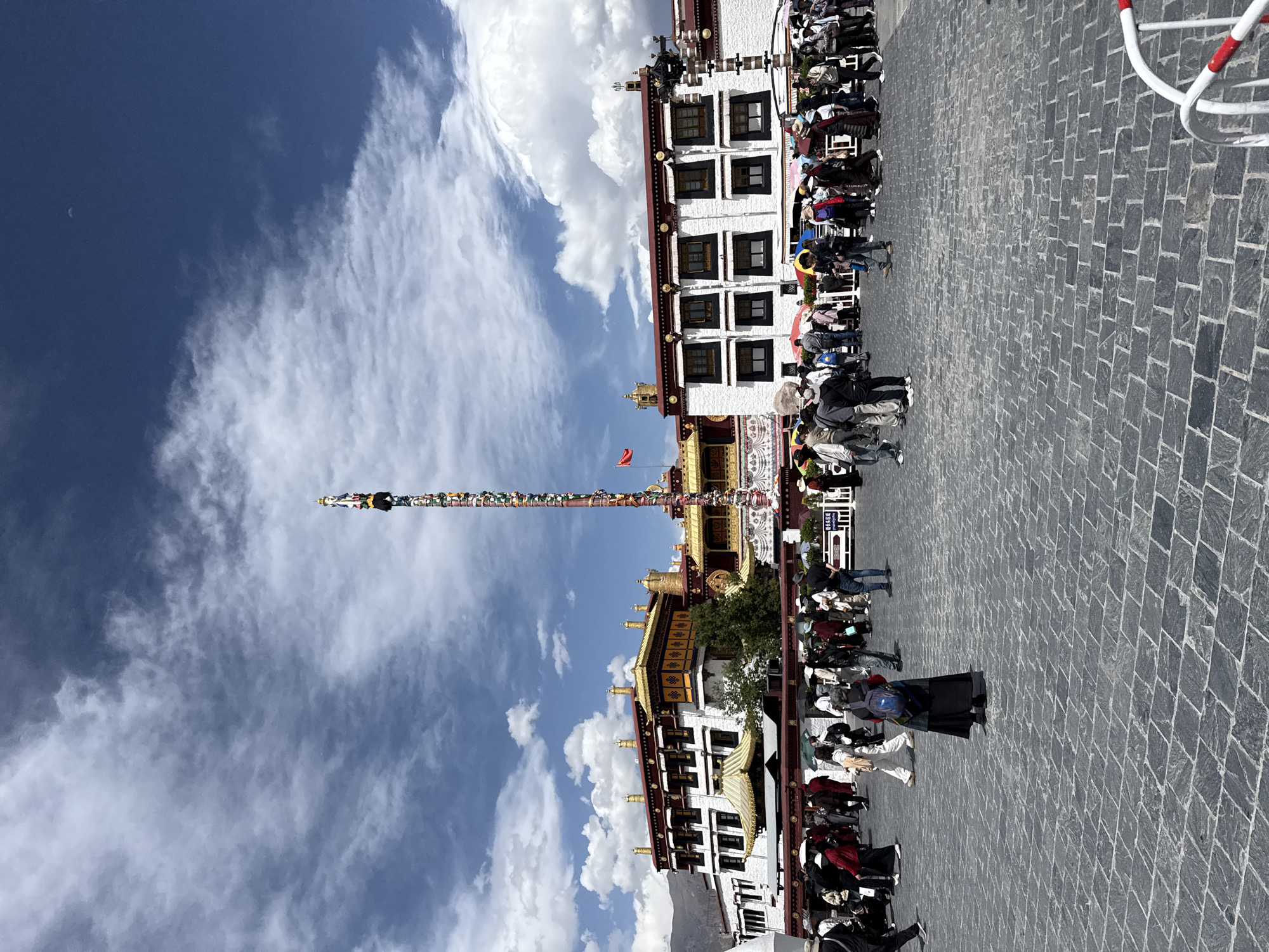 The image shows a crowd of people gathered in front of a traditional Tibetan building in the city of Chengguan District, China. The building has a white façade with dark windows and a gold-trimmed roof. A tall pole, adorned with prayer flags, rises in front of the building. The sky is blue with scattered white clouds. The ground is paved with gray bricks. The people are dressed in a variety of clothing, and some are wearing face masks.