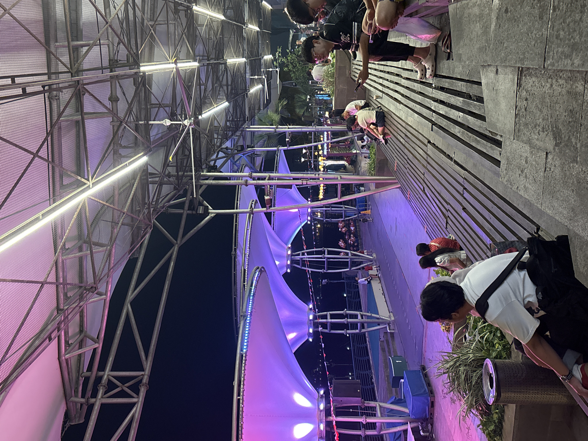The image depicts an outdoor seating area with a tiered wooden structure resembling bleachers, likely located in North Jakarta, Indonesia. The seating area is illuminated by purple and white lighting emanating from above, where a complex network of metal beams and large industrial fans are visible, suggesting a covered outdoor space. Several individuals are seated on the bleachers, some looking at their phones, others engaged in conversation. One person in the foreground is wearing a white t-shirt and black pants, with a black bag slung over their shoulder. The background reveals more of the outdoor venue, with a pathway, some greenery, and hints of buildings and lights in the distance, indicating an urban setting. The overall atmosphere appears to be one of relaxation and social gathering in a well-lit, modern outdoor space.